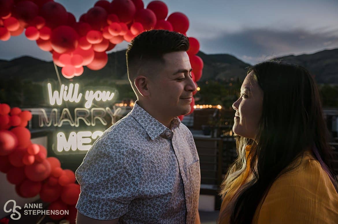 A newly engaged couple after sunset on a rooftop in Golden with the words "will you marry me?" lit up in neon behind them.