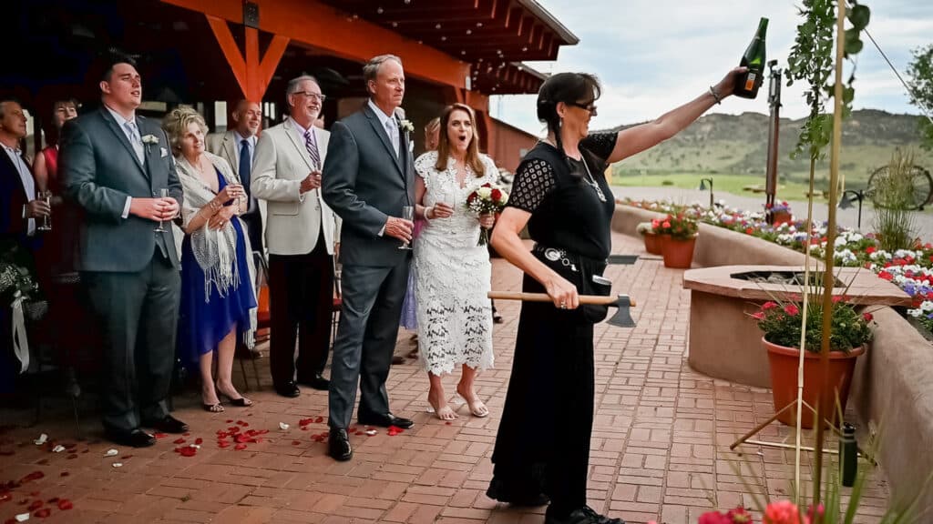 A woman dressed in black uses a tomahawk to open a bottle of champagne to the amazement of the wedding couple and their guests.