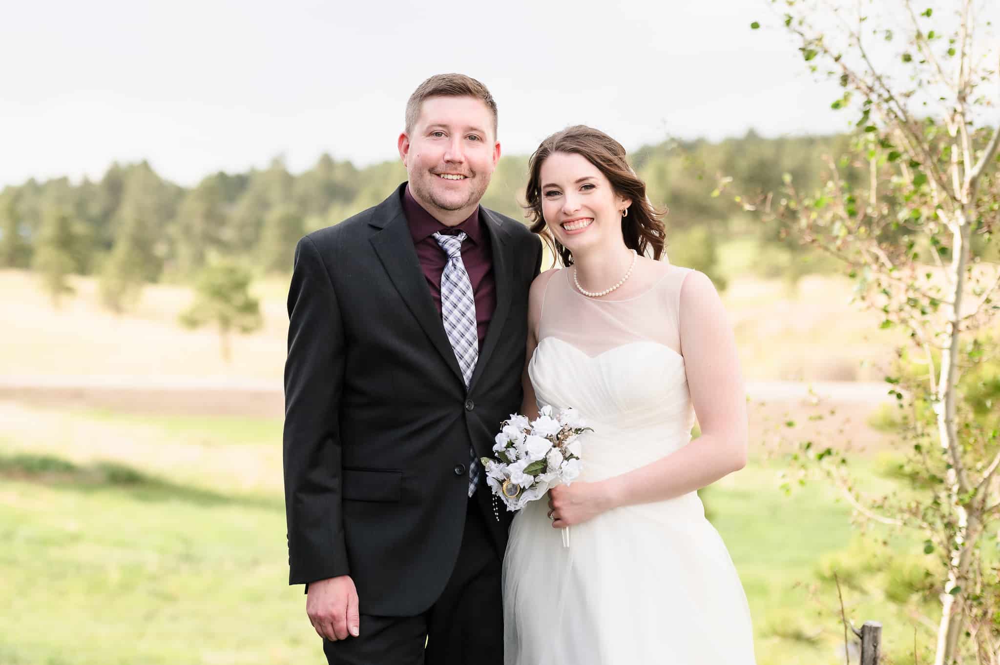 A bride and groom at the rustic 1C Barn in Monument, Colorado pose for a portrait in front of the natural grasses and trees.