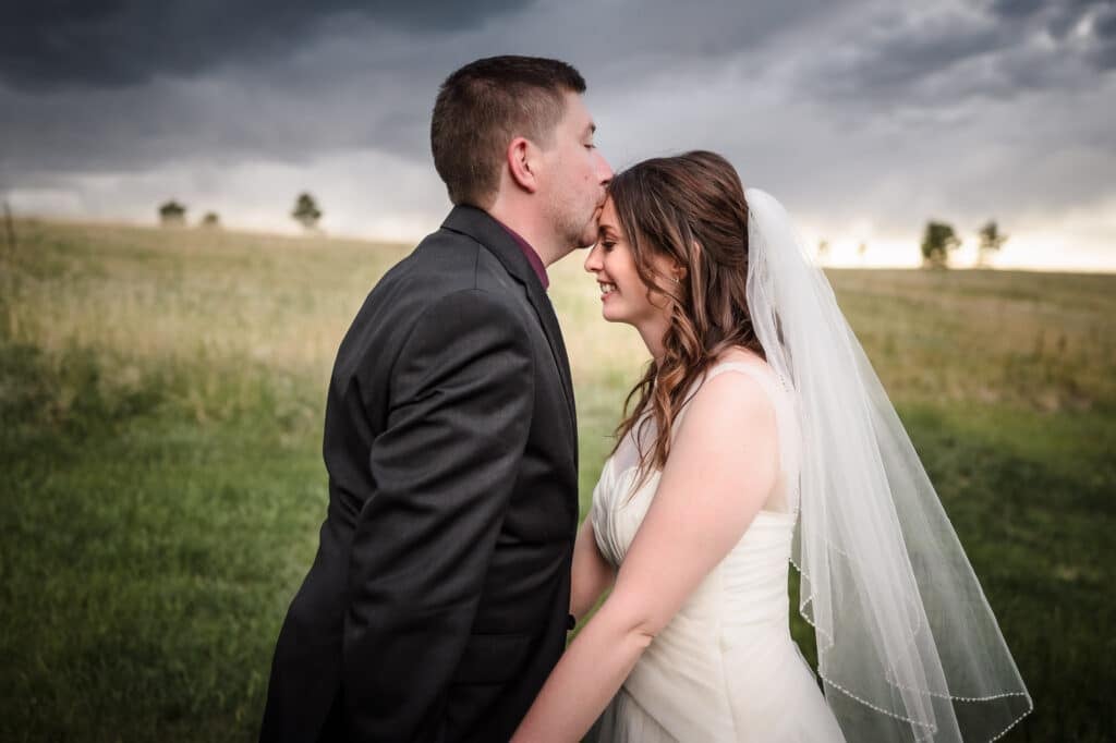 A groom kisses his bride on the forehead as the rain clouds start coming in over the prairie grass in Monument, Colorado.