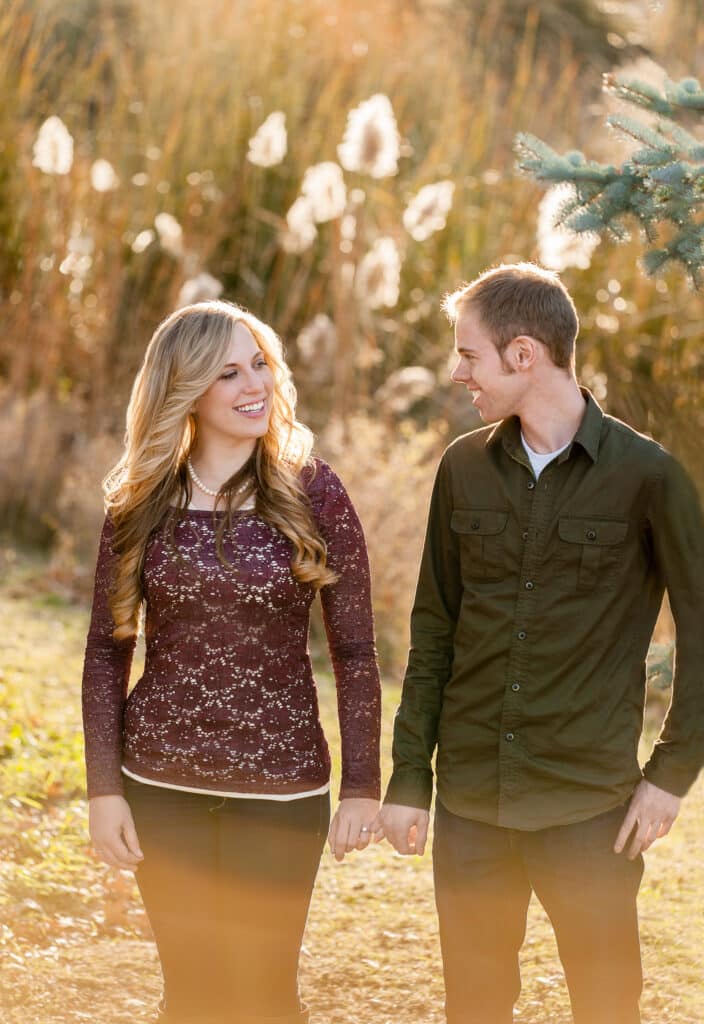 A man and woman hold hands as they walk in front of pompas grass illuminated with the sun.