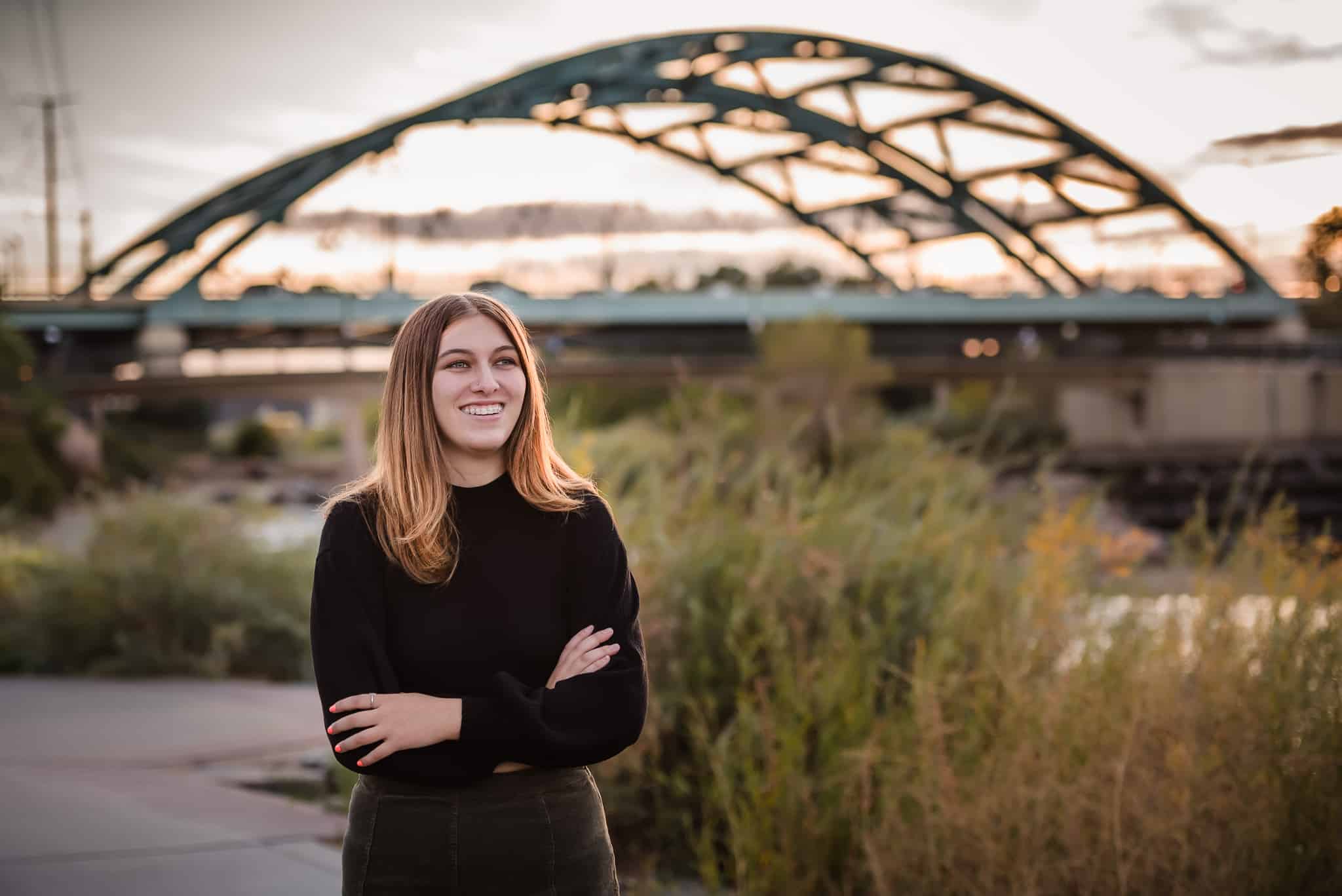 A young woman crosses her arms and stands in front of an arching bridge as the sun sets in downtown Denver