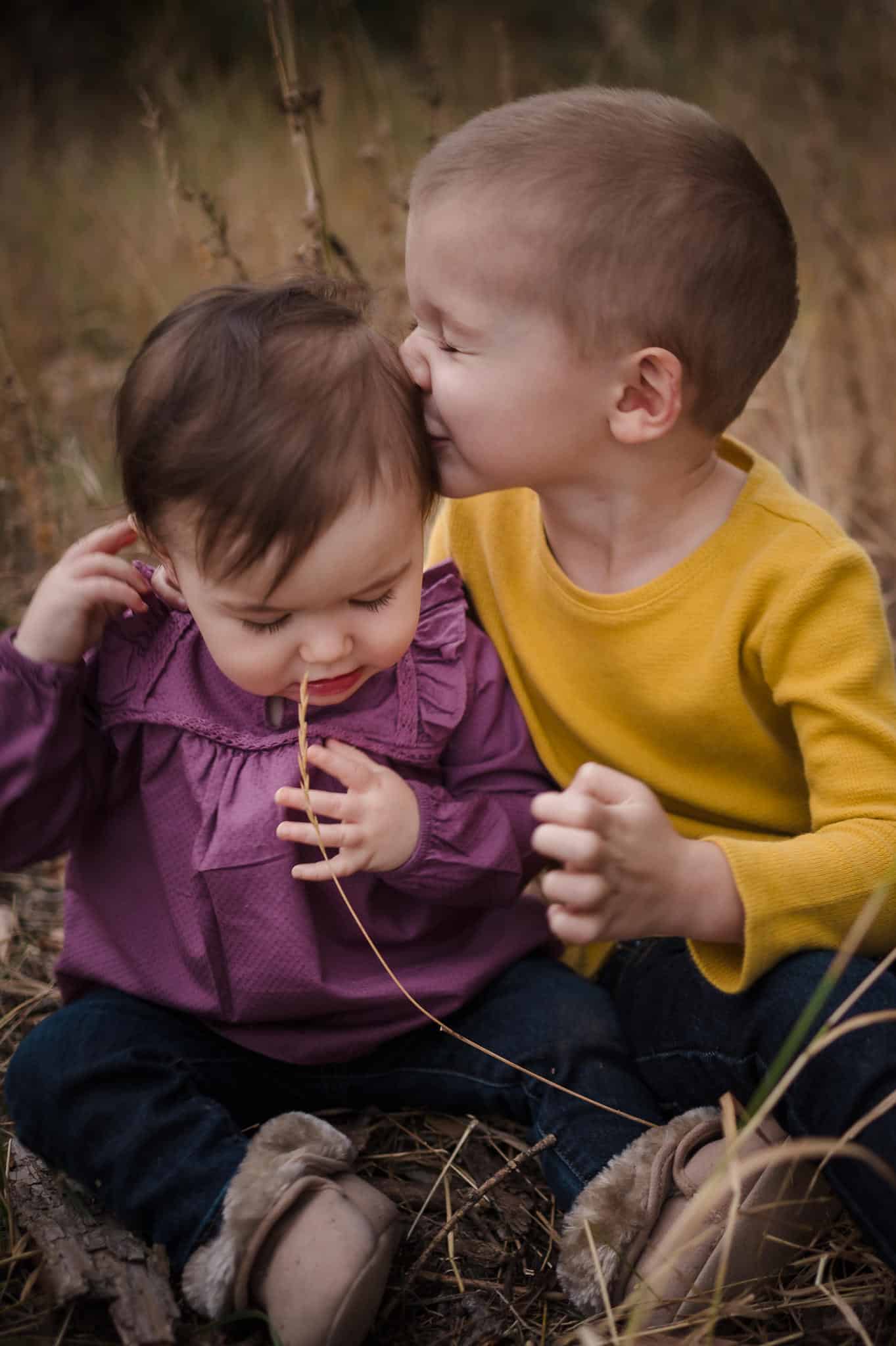 A young boy awkwardly kisses his toddler sister as they sit in the grass and play together.