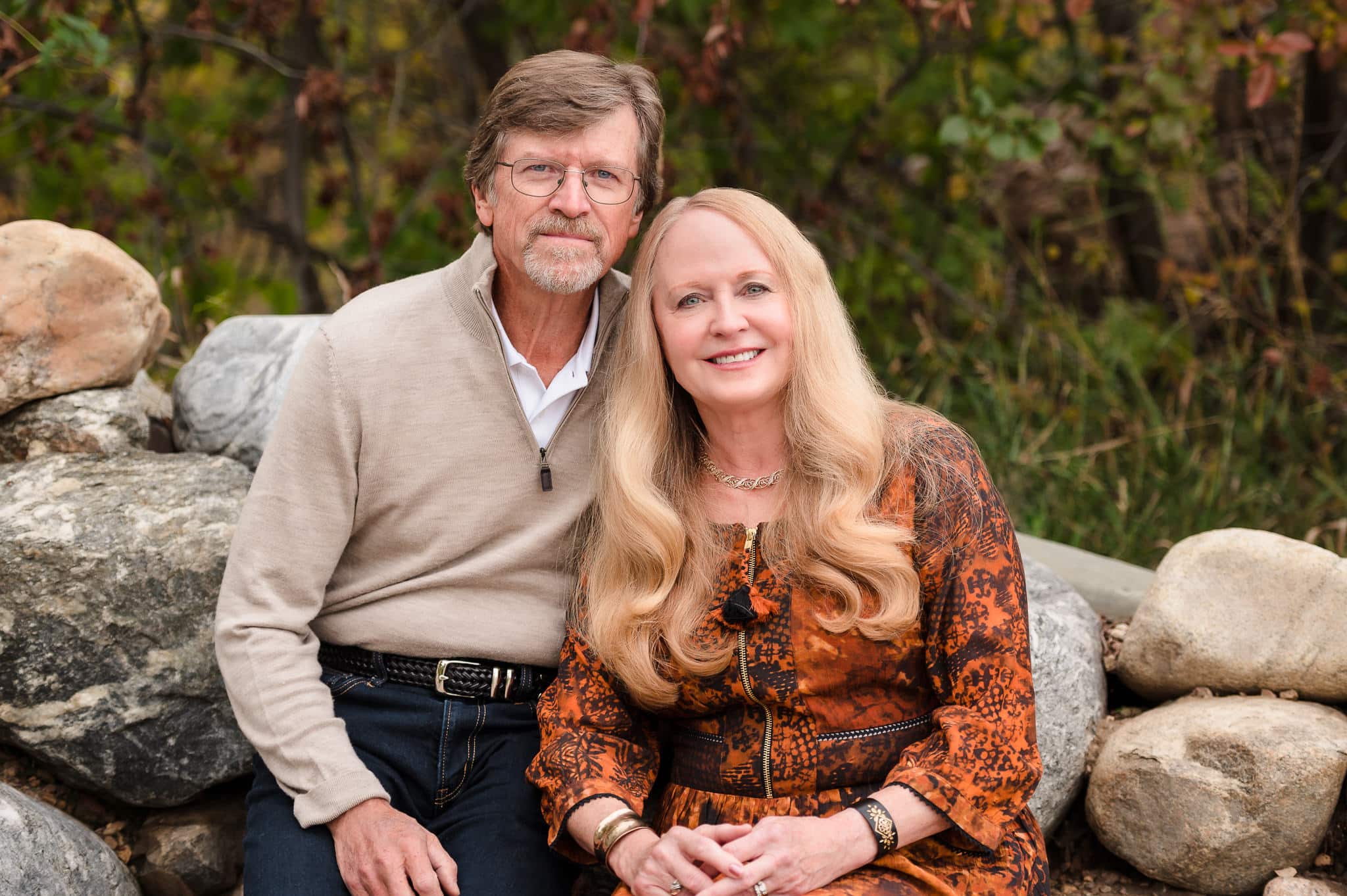 A man and woman in beige and burnt orange colored attire sit on a rocky edge at the Sweetheart Winery in Loveland, Colorado.