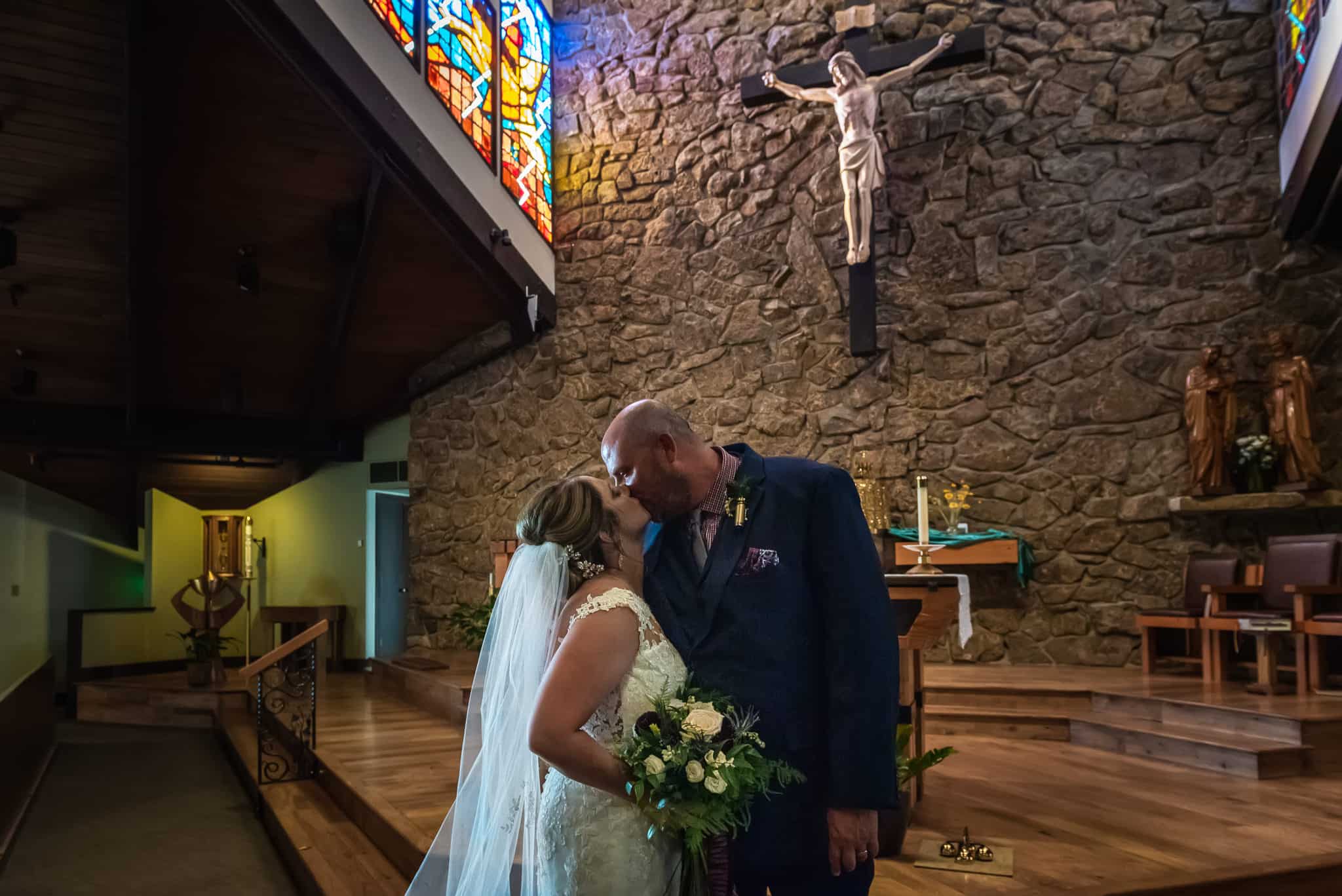 Natural light photo of a bride and groom sharing a kiss at the altar after their ceremony in a Catholic Church.