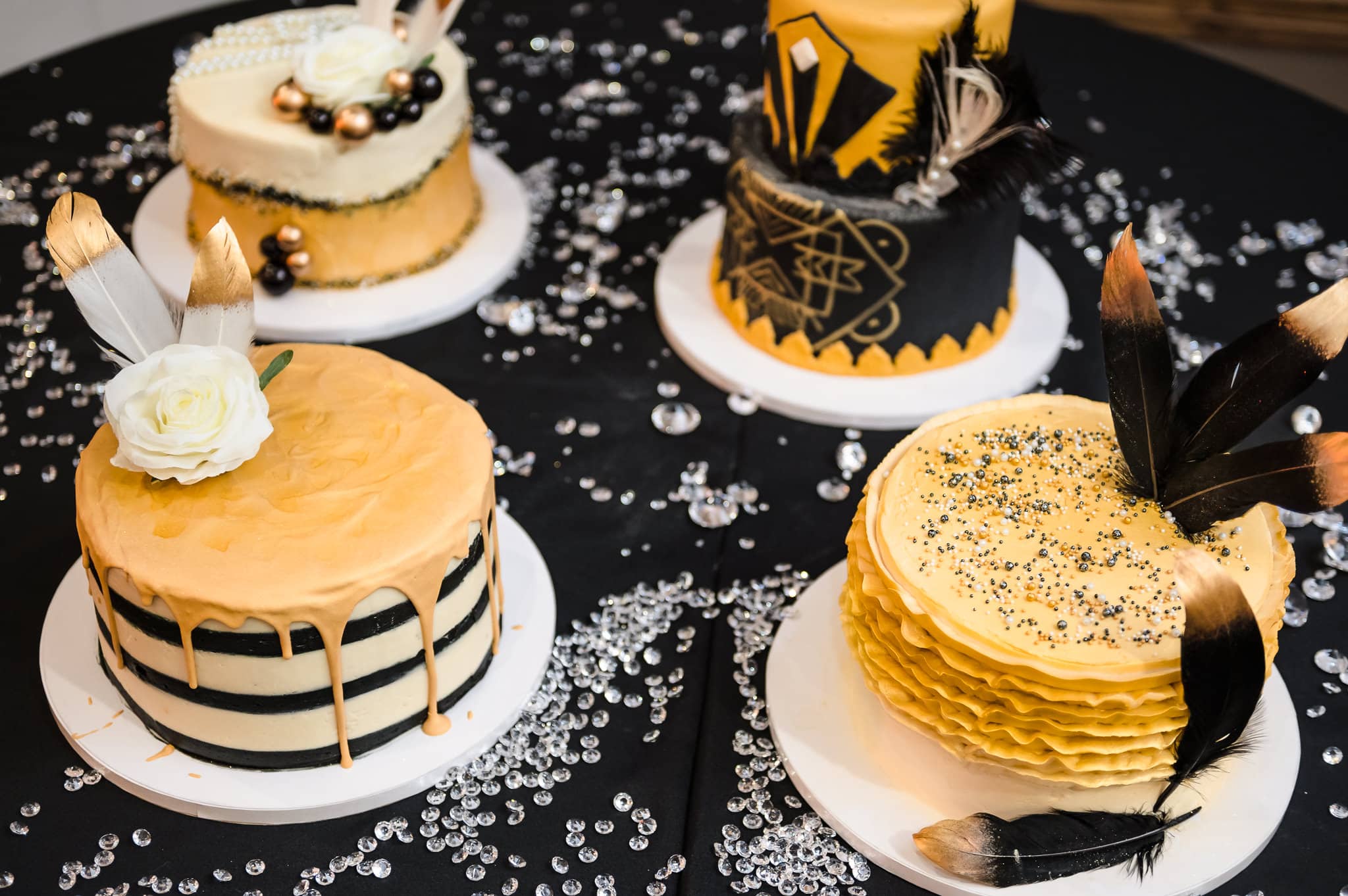 Four art-deco decorated cakes on a table with faux diamonds feature candy beads, feathers, and fondant in gold and black. 