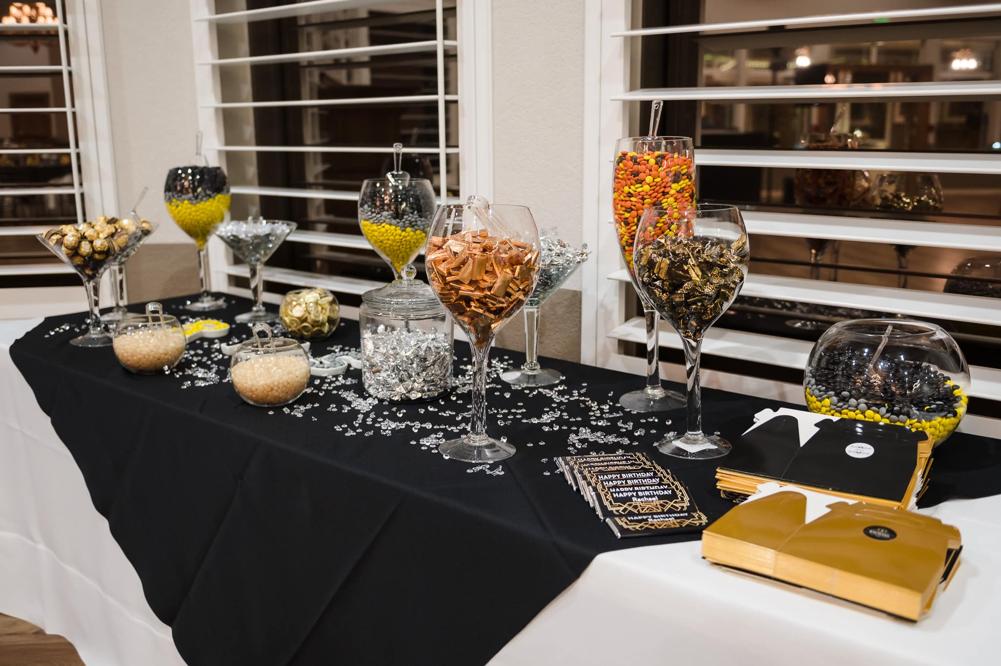 Plentiful amounts of clear glass candy jars in gold and black on a table ready for guests at a 40th birthday Great-Gatsby themed surprise party.