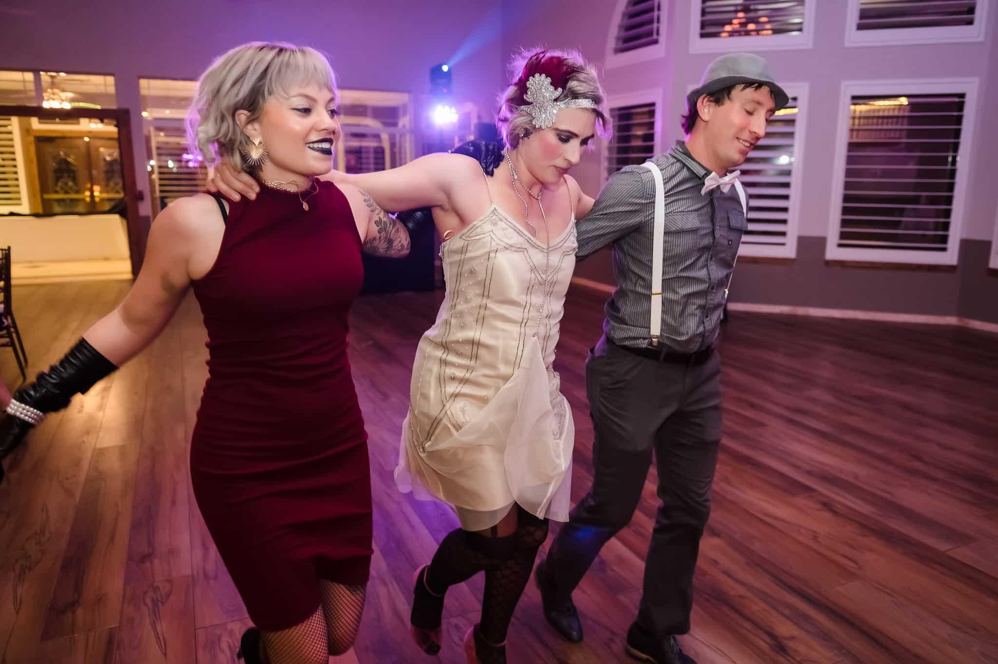 Two woman and a man dressed in 1920s inspired attire link arms and dance in a circle around the specially lighted room to Cotton Eye Joe at a Great Gatsby themed birthday party.