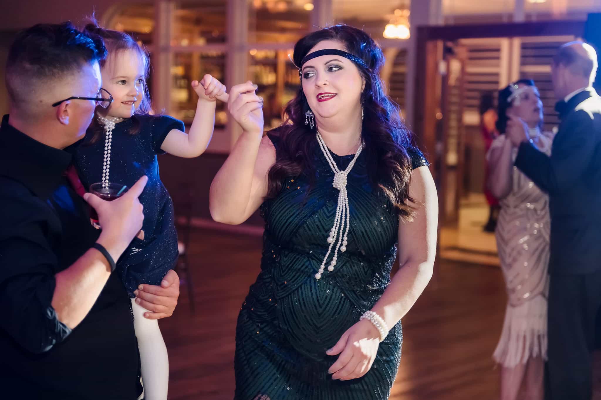 A man lifts his younger daughter to dance with him and his wife at a 1920s Great Gatsby party.