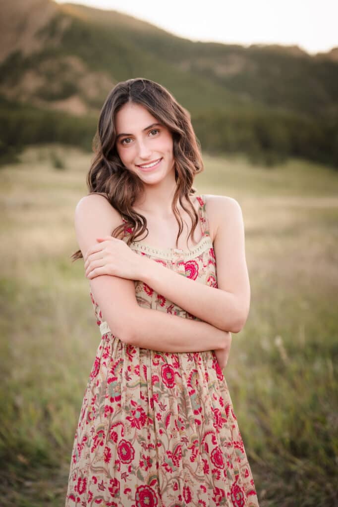 Best high school senior portraits for young women at Chautauqua Park, Boulder. A young woman standing in prairies grass in a long flowing red dress with her arms gently crossed and the Flat Irons in the background