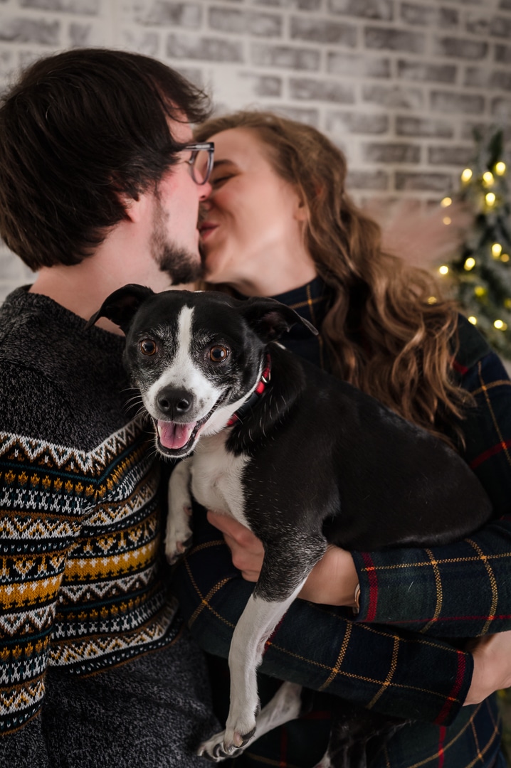 A young millennial couple kiss while their dog looks into the camera.