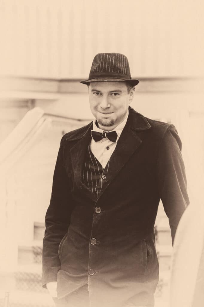 A male guest wearing a bowler hat, tie, and vestcoat for a 1920s Great Gatsby birthday party poses with a subtle smile.