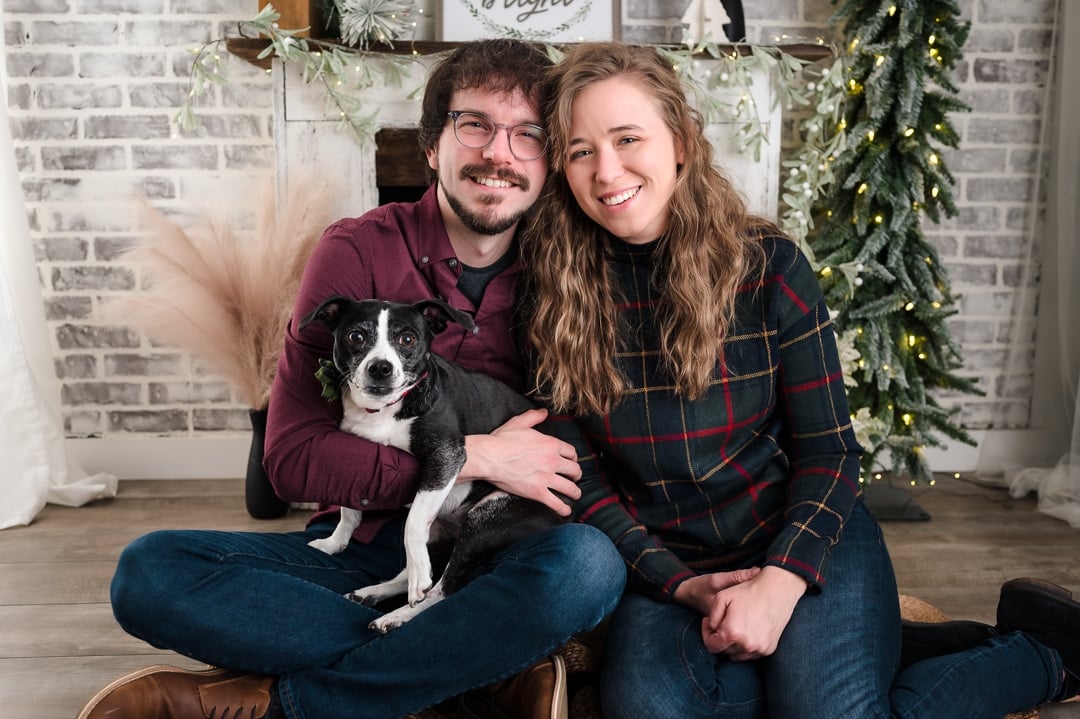 A young millennial couple and their puppy Zelda sit in front of a holiday decorated fireplace and mantel during a holiday photo session.