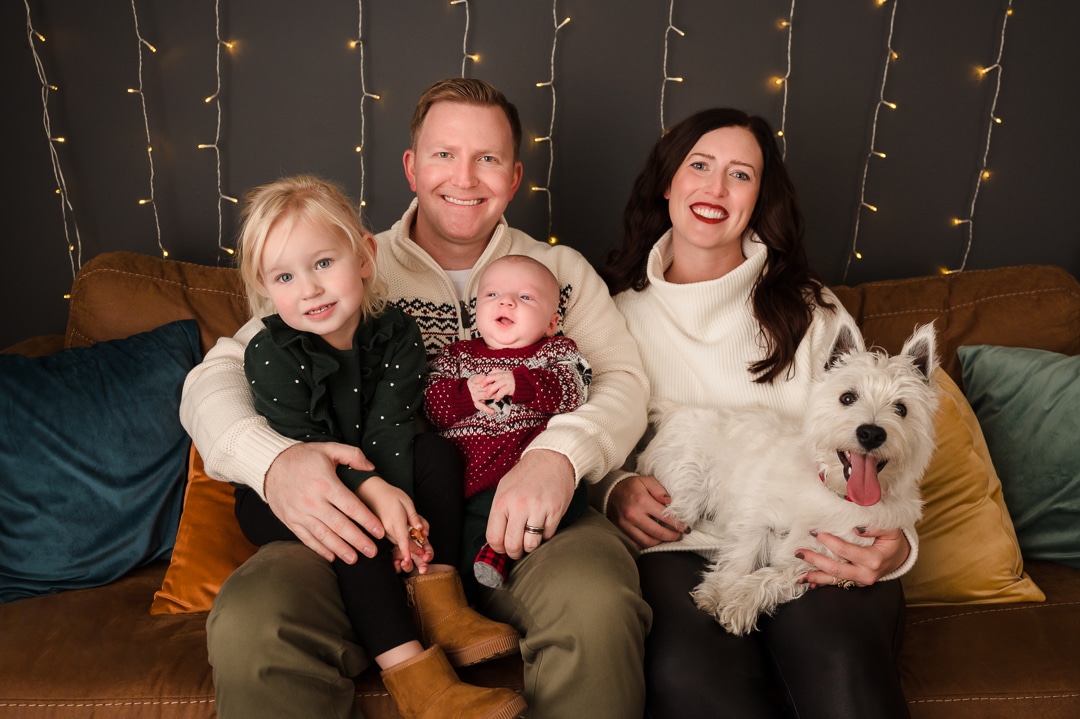 A mom, dad, young girl, and a newborn boy sit with their dog on a couch during a family photo session.