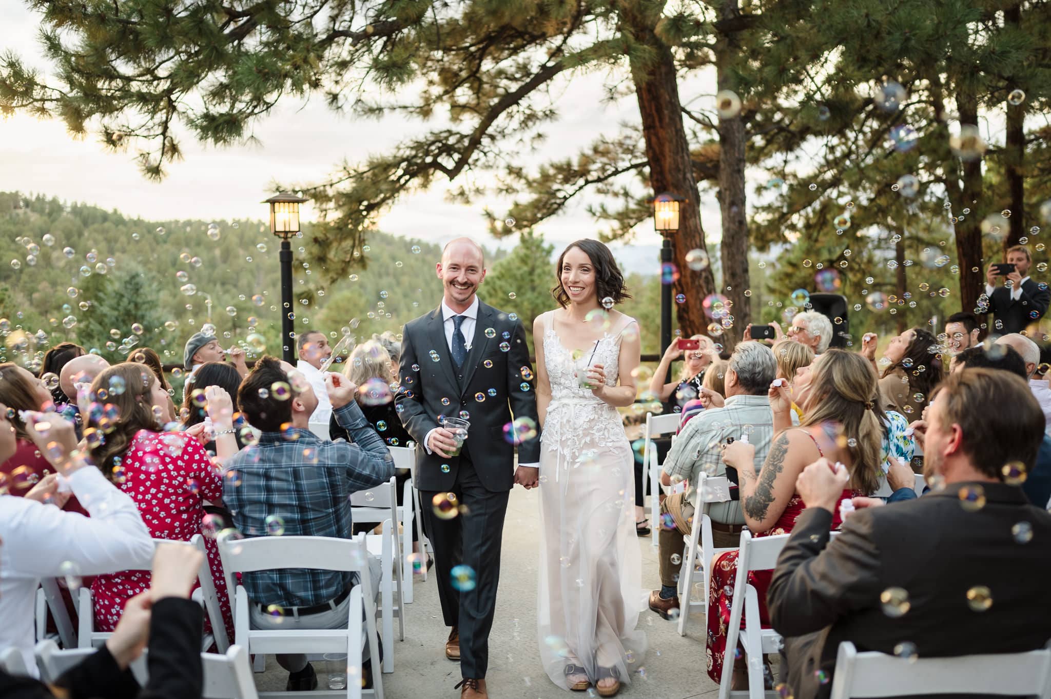 A man in a suit and woman in a wedding dress depart their vow renewal ceremony down the aisle as guests blow bubbles in celebration.