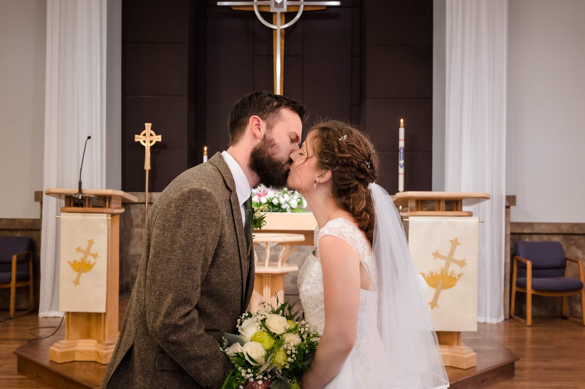 The bride and groom lean over the bridal bouquet and kiss at the altar of the church.