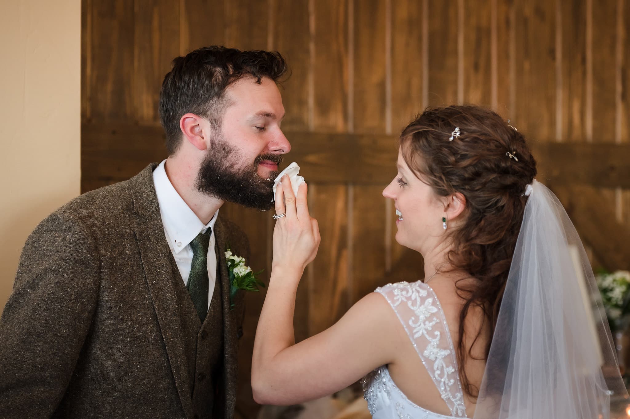 The bride wipes crumbs and frosting from the beard of her husband following the traditional cake smash at their wedding.