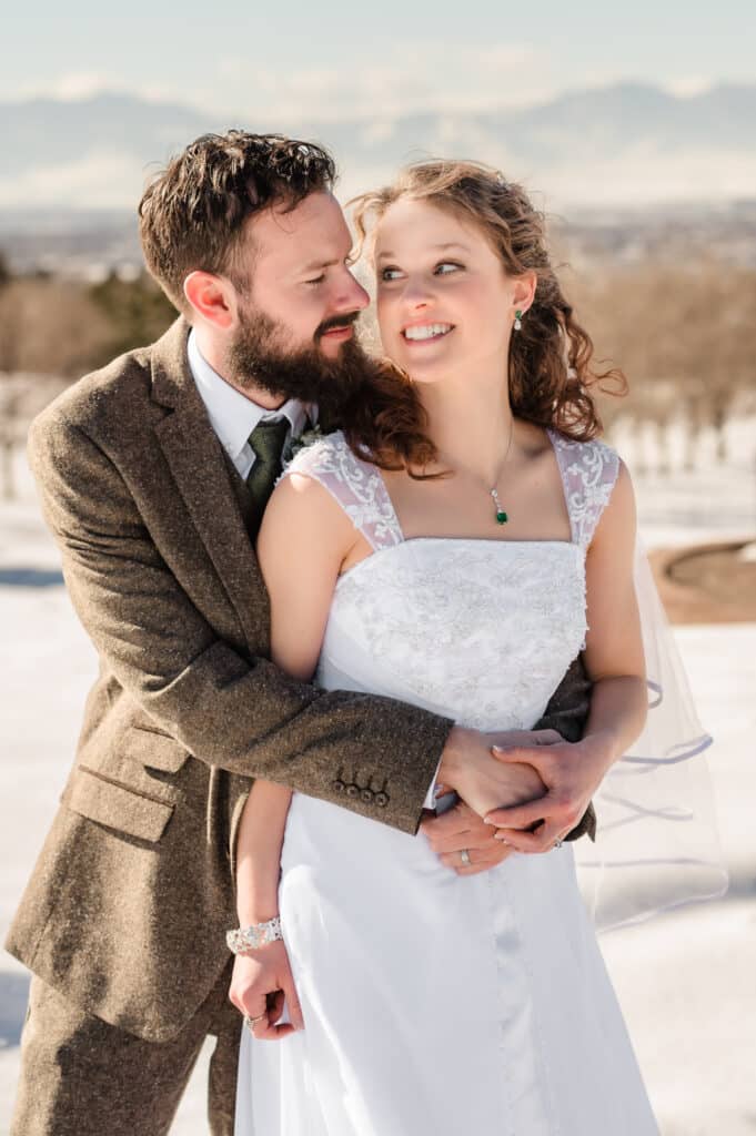 The wedding couple smile at each other in a snowy scene with the mountains in the background.