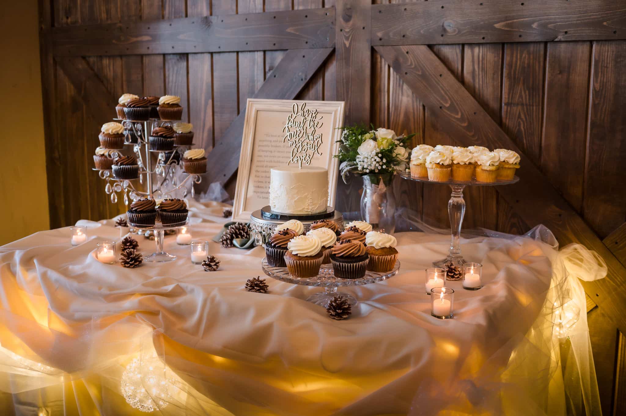 Wedding dessert table filled with crystal pedestal stands of cupcakes and a miniature white fondant wedding cake with mountain details in frostingin front of a dark brown rustic wooden paneled barn style door features warm glowing holiday lights around the edges.
