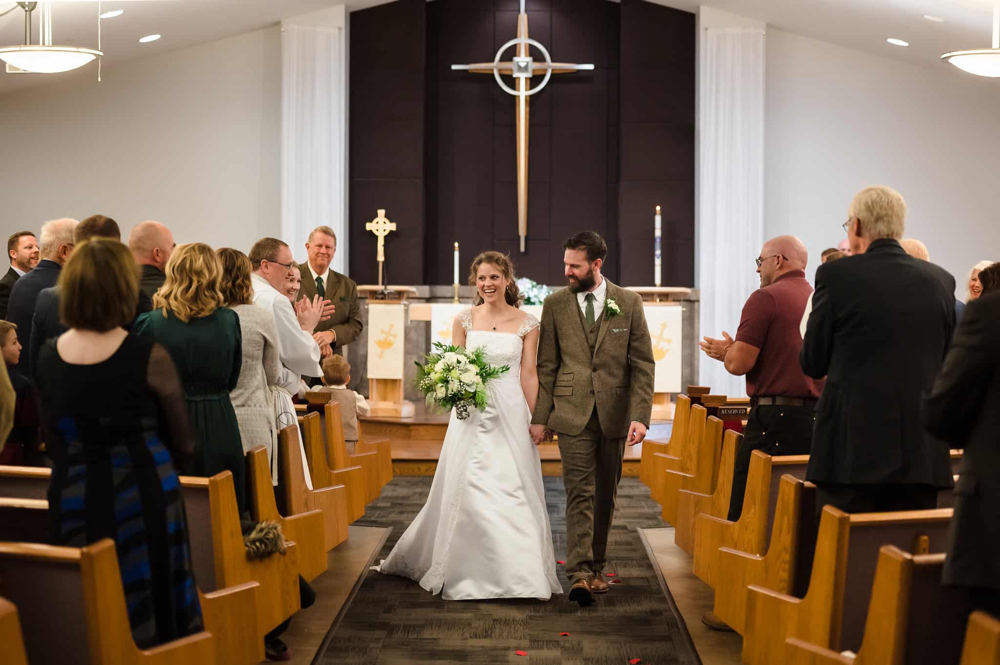 The ceremony completed, the wedding couple hold hands and walk down the aisle of the church between the pews.