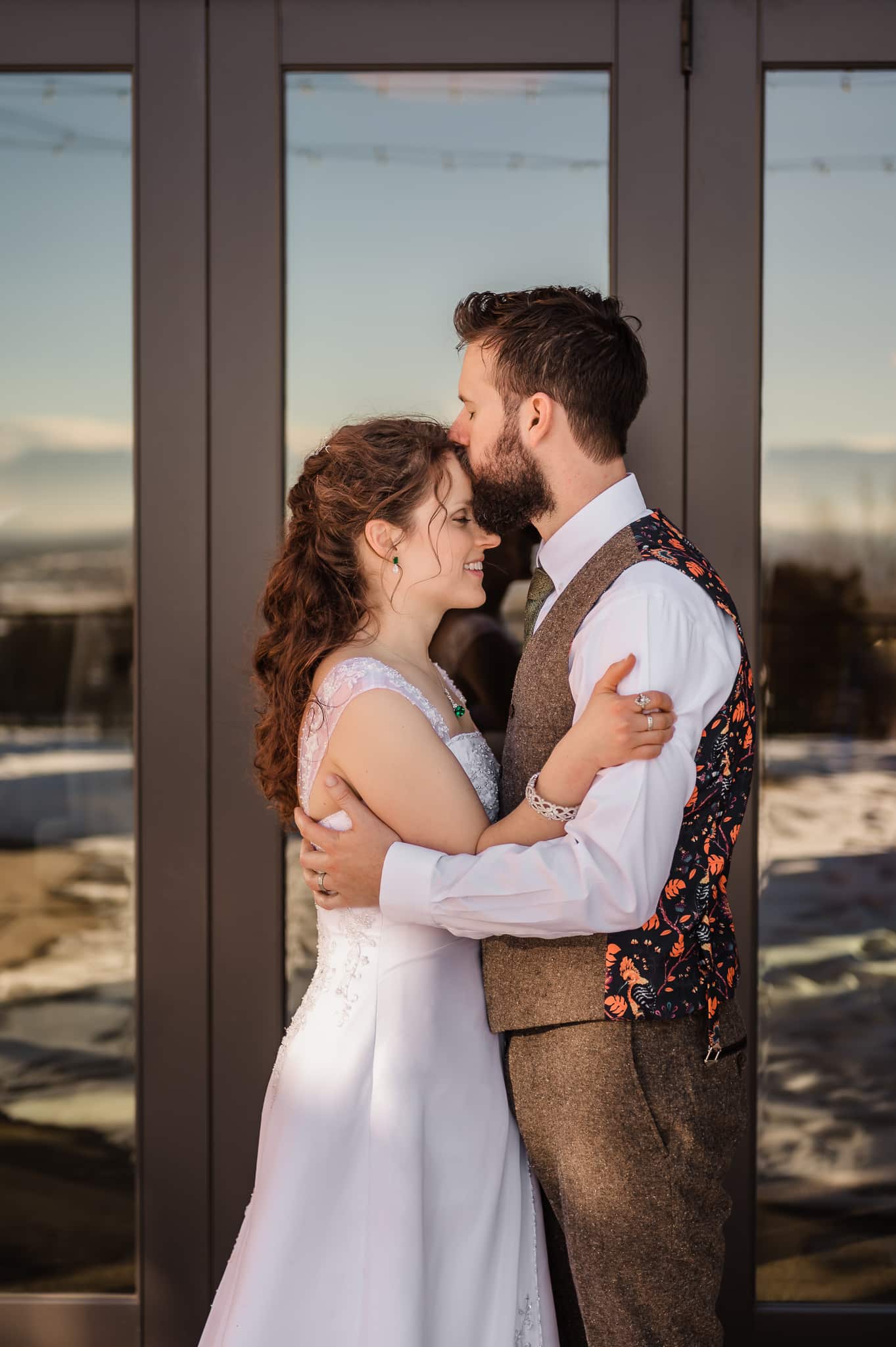The bride and groom face and hold each other in front of reflective doors that mirror the mountains and warm light across the way to the west.