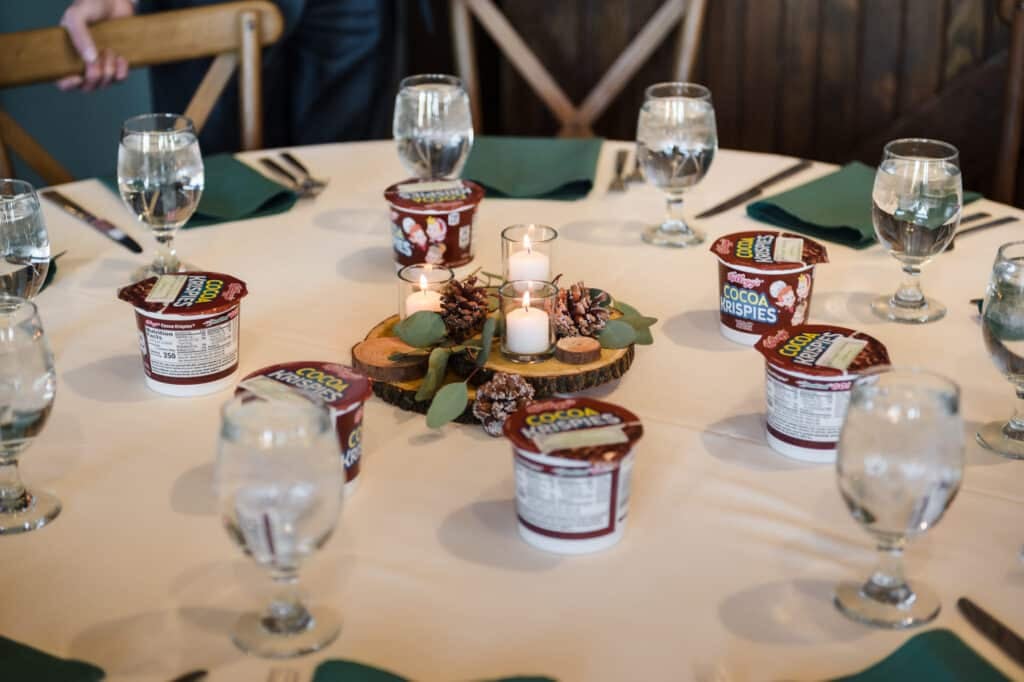 The table settings at this wedding included candles, pinecones, and leaves on a wooden centerpiece, water glasses, green napkins, and single-serve containers of Cocoa Crispies brand cereal.