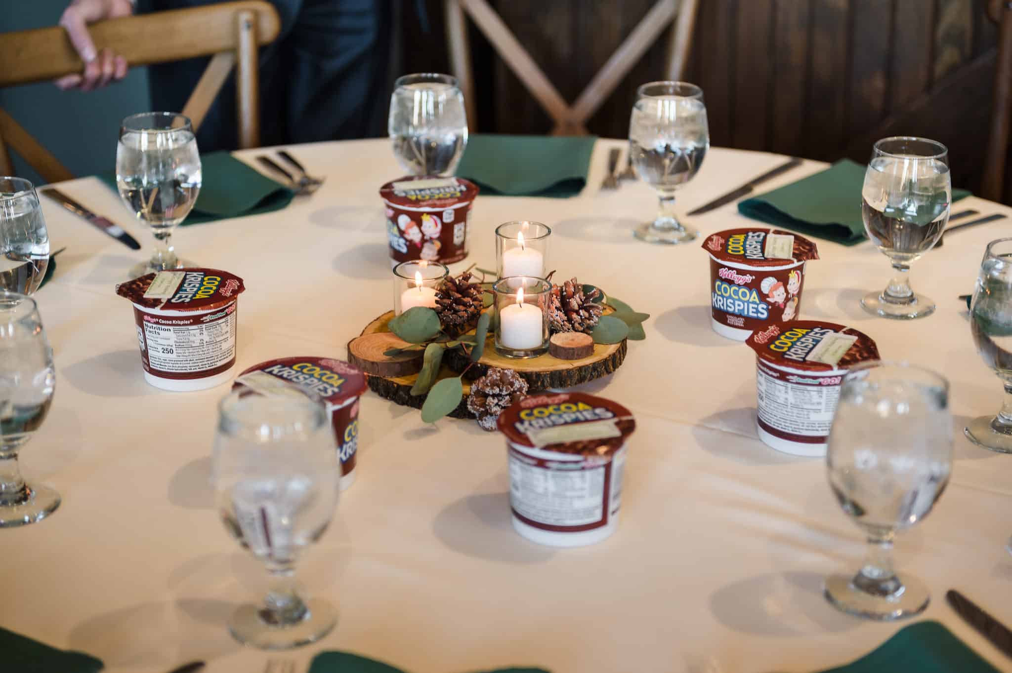 The warm and earthy colors of brown and green at this wedding table make a memorable winter wedding color scheme. Included are ivory candles, pinecones, and leaves on a wooden centerpiece, water glasses, green napkins, and single-serve containers of Cocoa Crispies brand cereal.