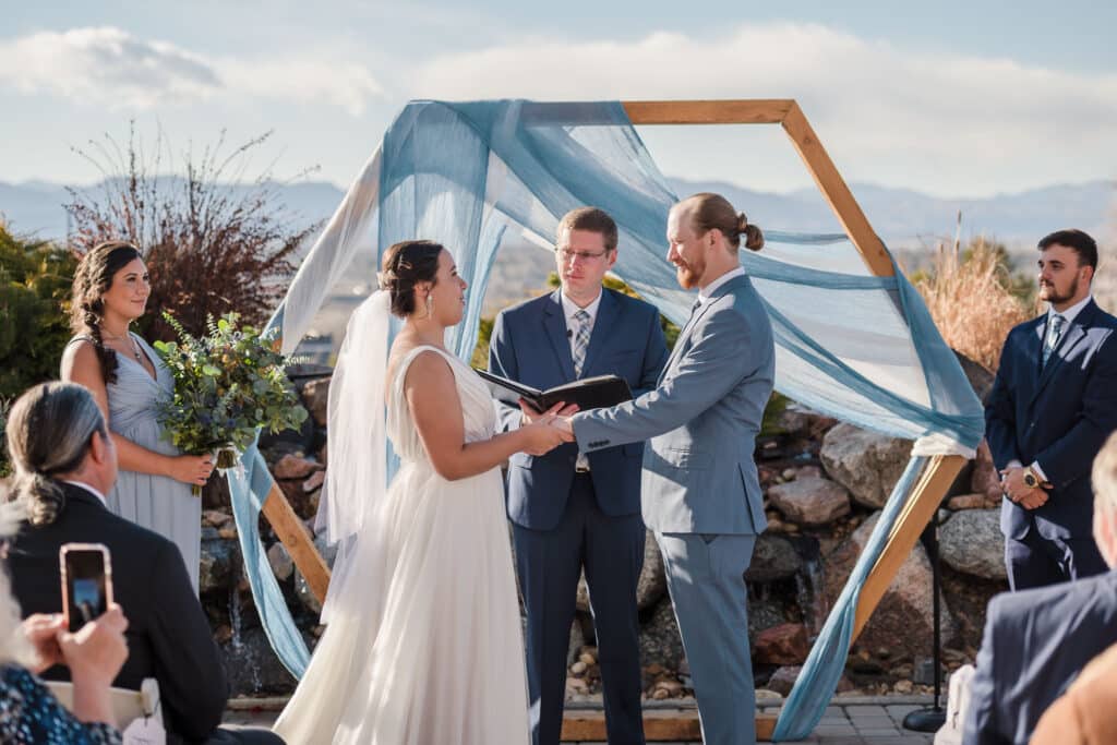A man and woman hold hands together under an archway draped with their dusty blue wedding colors at an outdoor spring wedding held at Ashley Ridge.