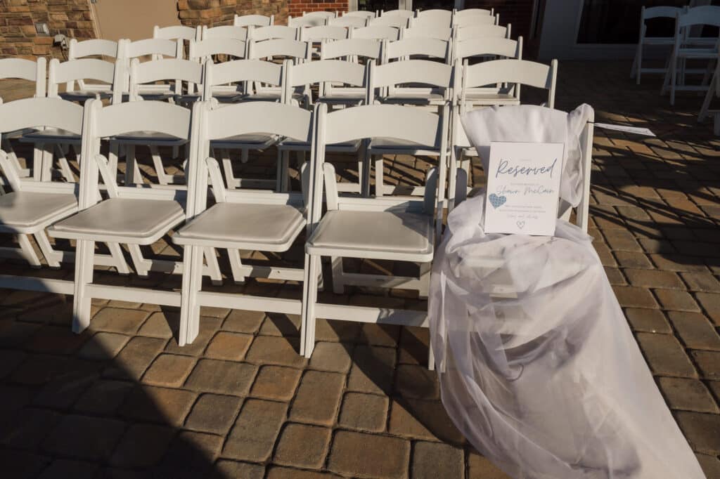 Rows of white chairs set up for a wedding. The farthest chair on the right is wrapped with fabric and bears a sign stating that it is reserved in memory of the groom's father.