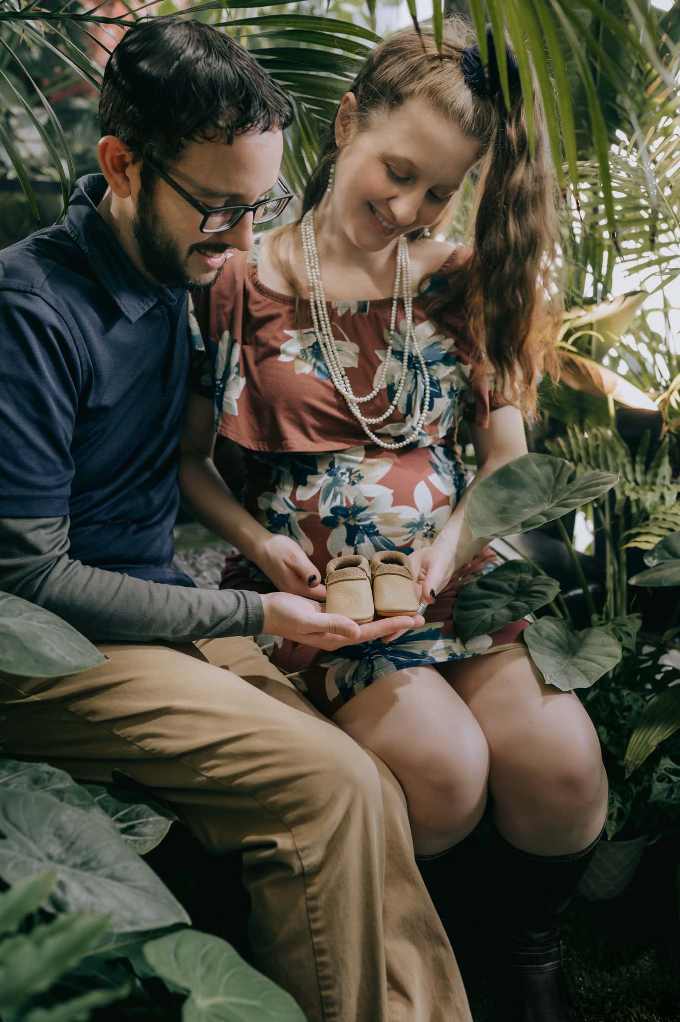 Sitting among jungle leaves, an expecting couple holds tiny baby booties.