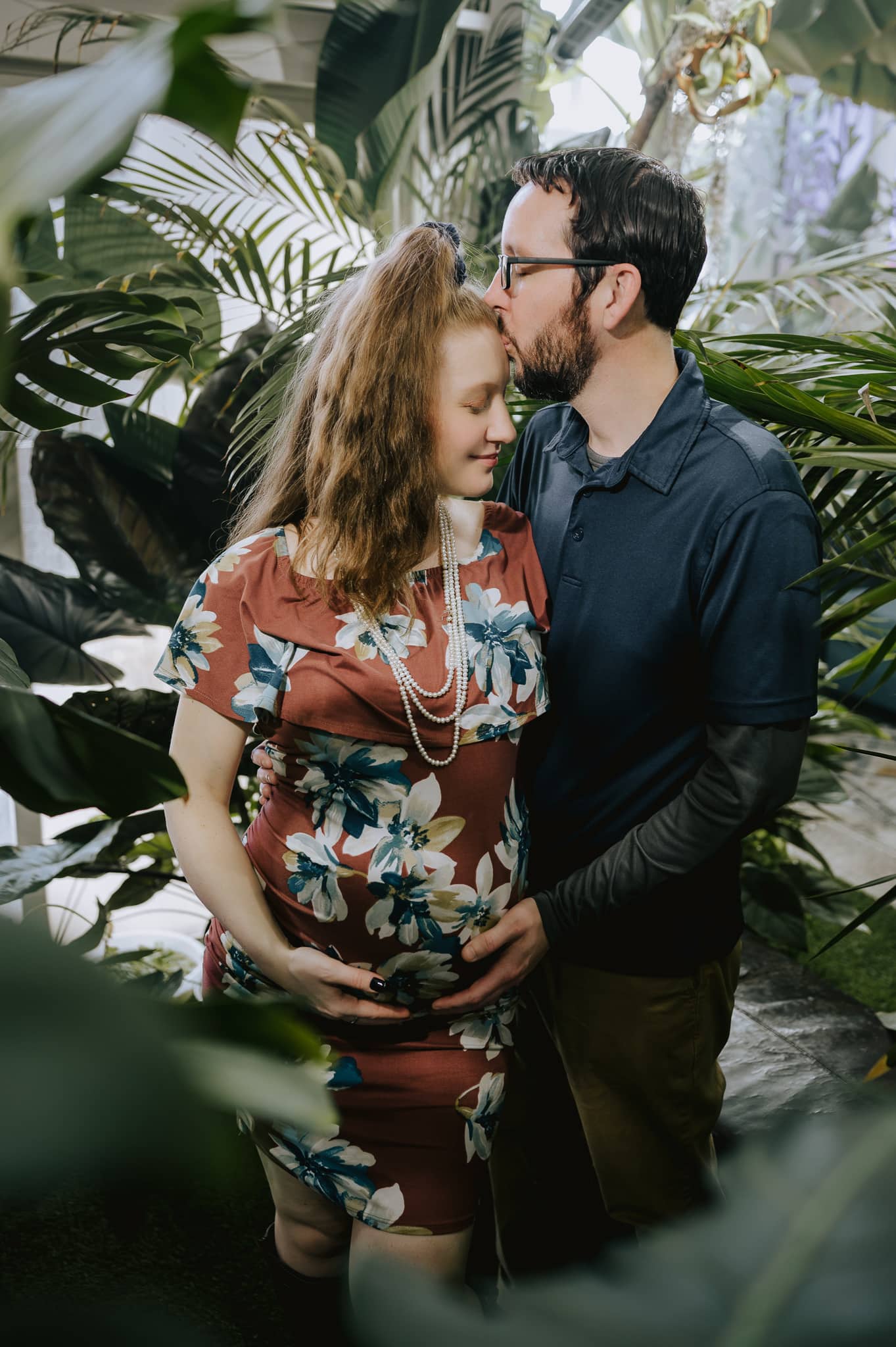 A husband kisses his expectant wife on the forehead during their unique jungle indoor maternity photo session.