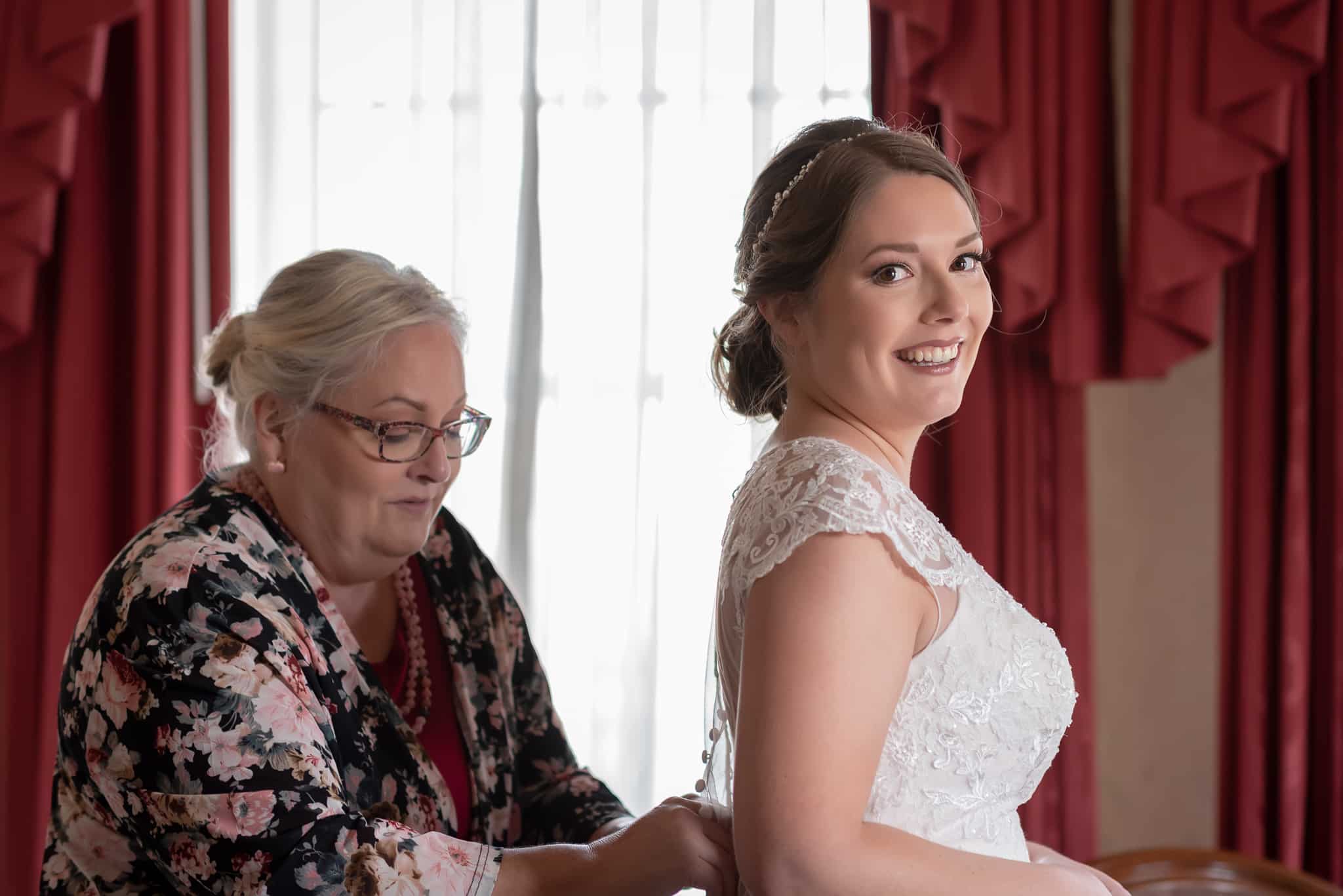 A mother honors her daughter at her wedding by helping with the small buttons on the back of her daughter's bridal dress.