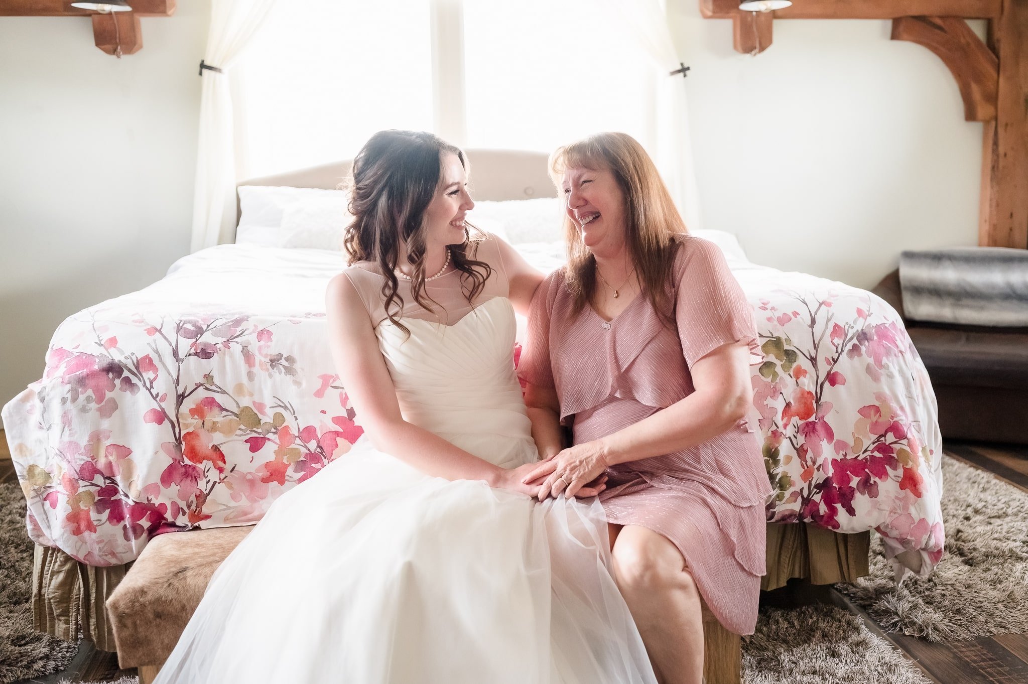 The mother of the bride laughs with her daughter as they sit on the edge of a bench in a bedroom.