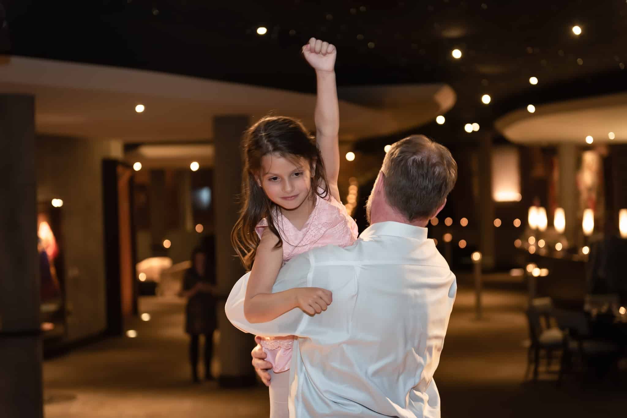 A young girl dances with her mother's husband during the wedding reception and raises her fist in victory.