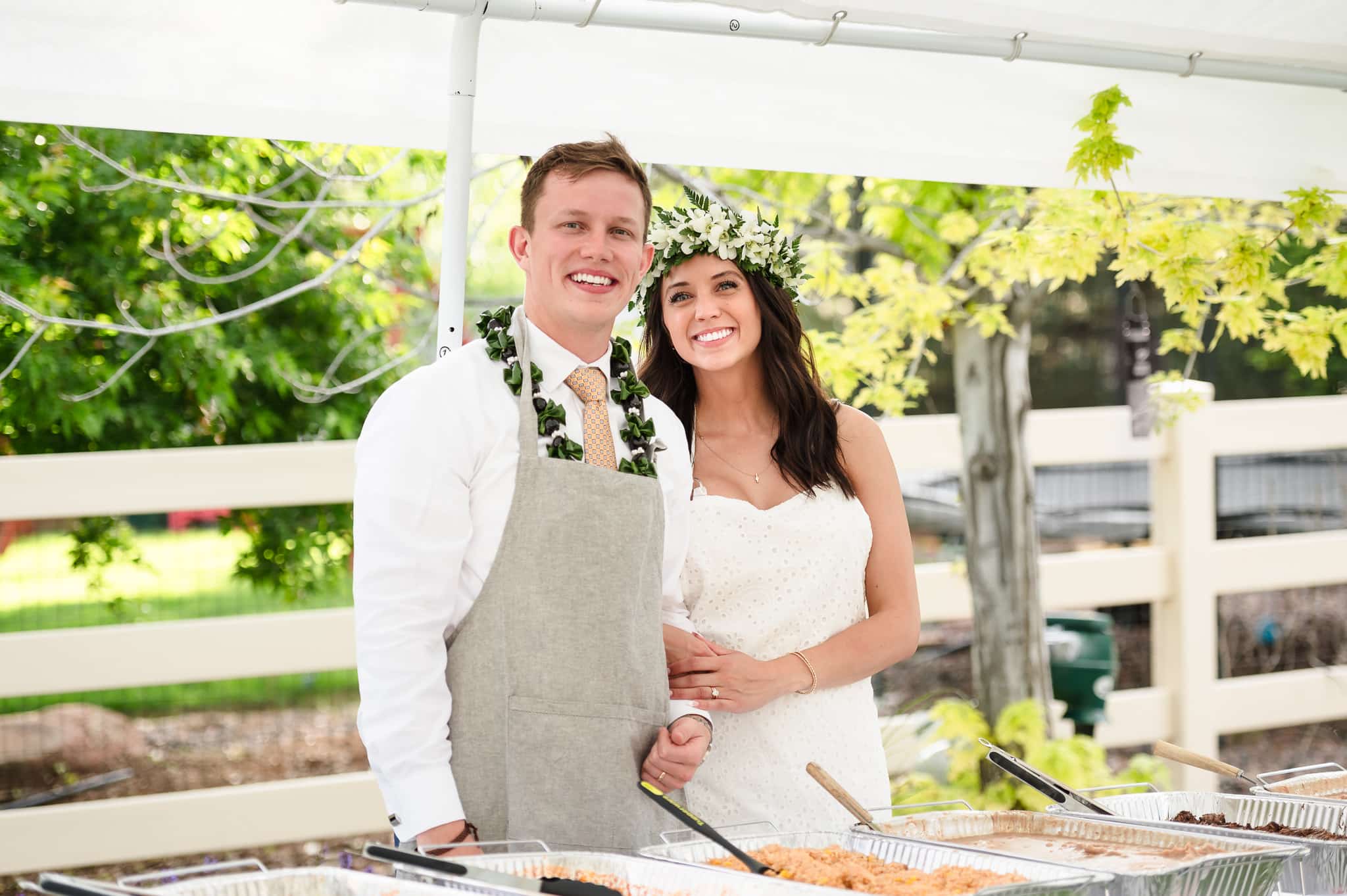 The bride and groom wear aprons over their wedding attire and are ready to serve their guests food from their buffet.