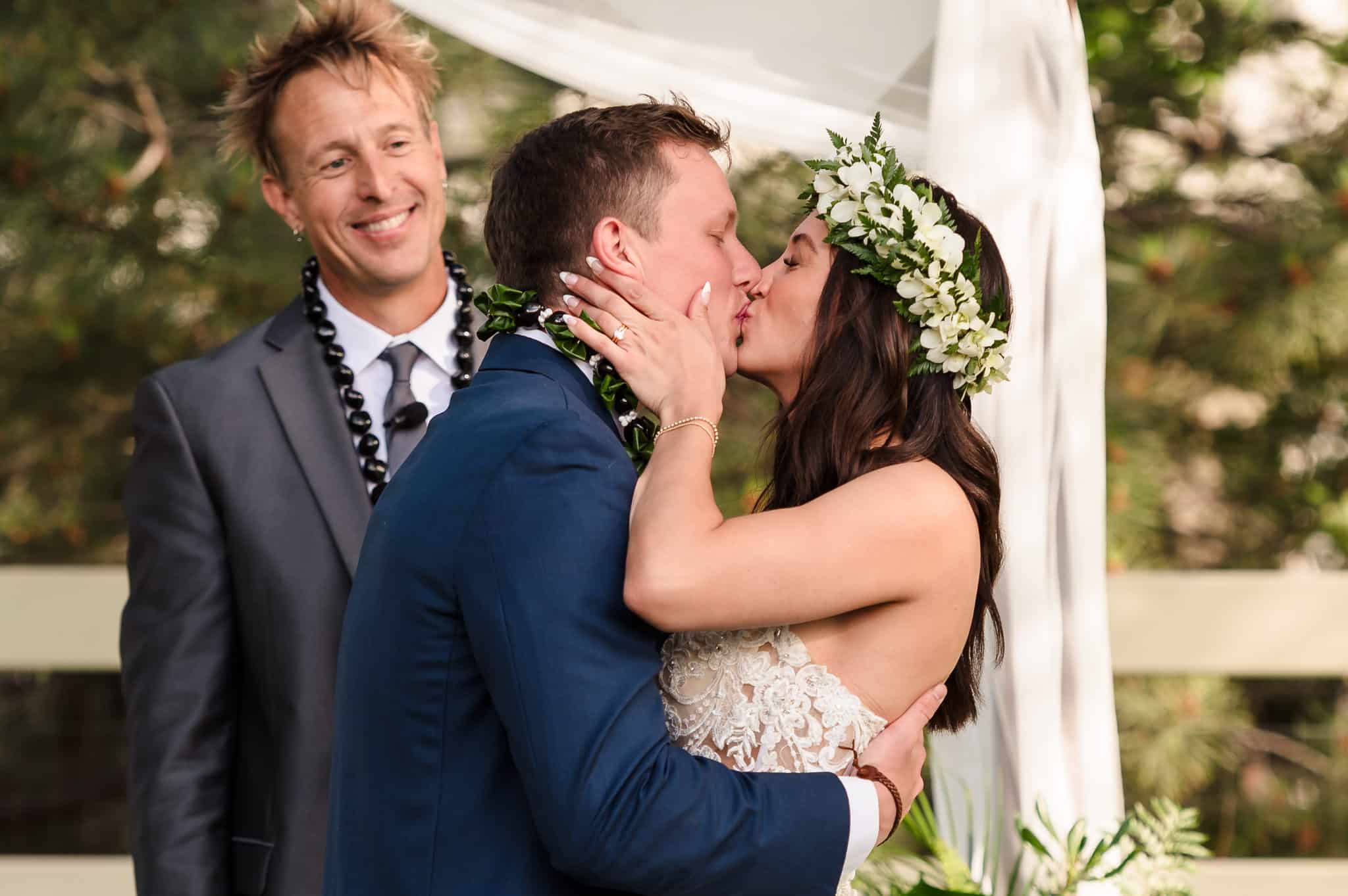 A smiling officiant looks on as the bride and groom share their first kiss at their Hawaiian-inspired backyard wedding.