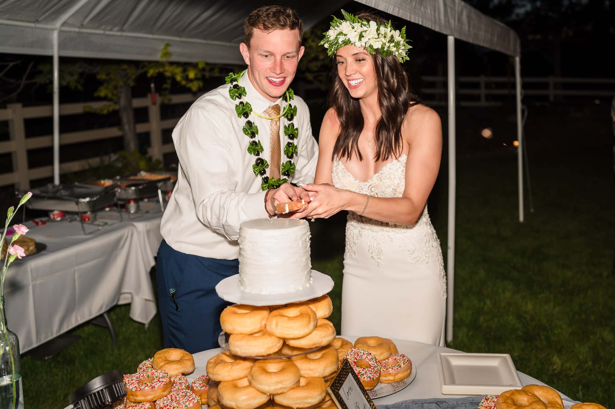 The bride and groom cut the wedding cake located behind the stacks of donuts.