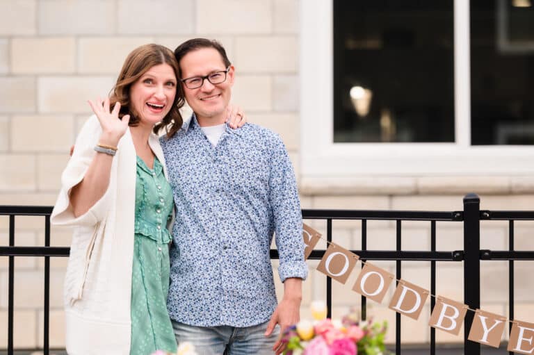 A wife and husband wrap arms around each other as the wife waves in farewell in front of the goodbye sign at their going away party.