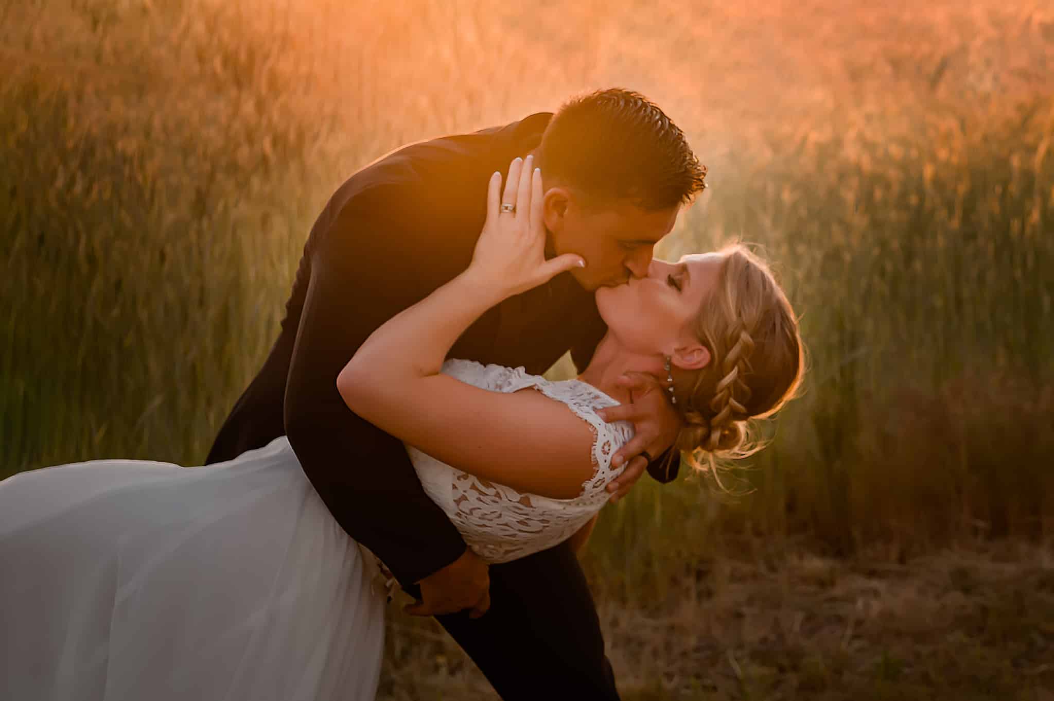 The groom dips the bride in the prairie grass as the setting sun gives a soft lens flare to the scene.