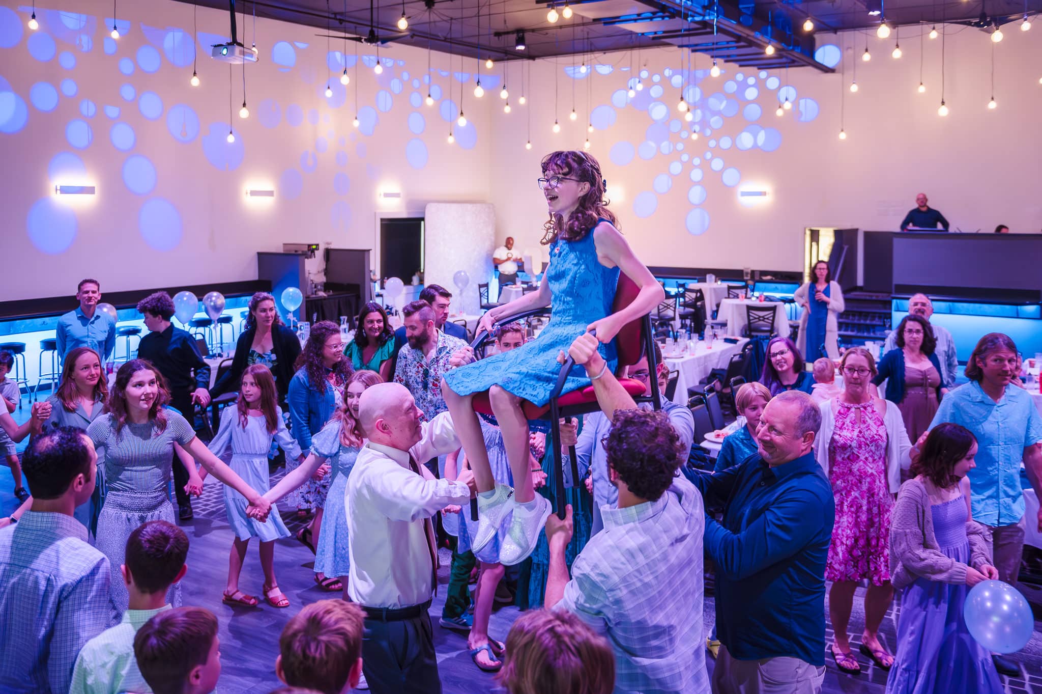 Side view of a young woman being lifted up in a chair during The Hora at her bat mitzvah.