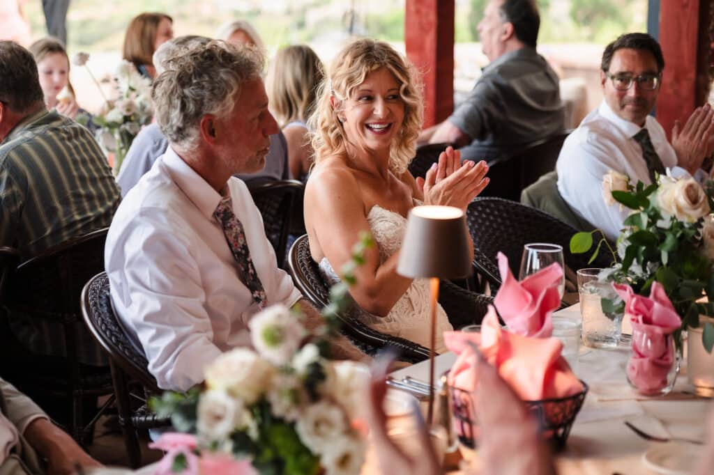 The bride, seated at a table next to her husband, claps after the words of welcome given by the officiant.