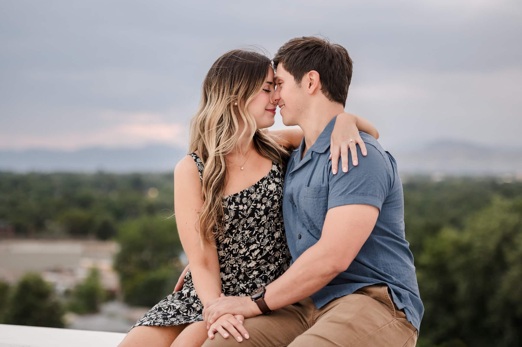 A couple sits on a wall at a park with the mountains blurred in the background and snuggles together after their engagement.