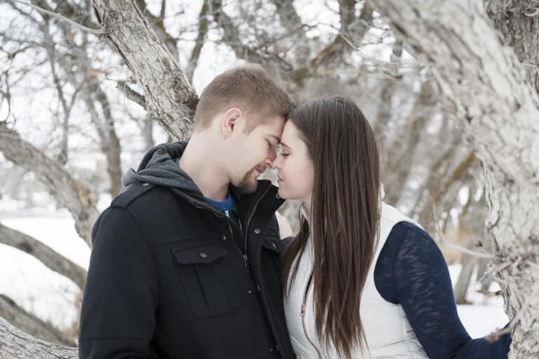 A man and woman snuggle together in front of a tree during an outdoor winter engagement session.