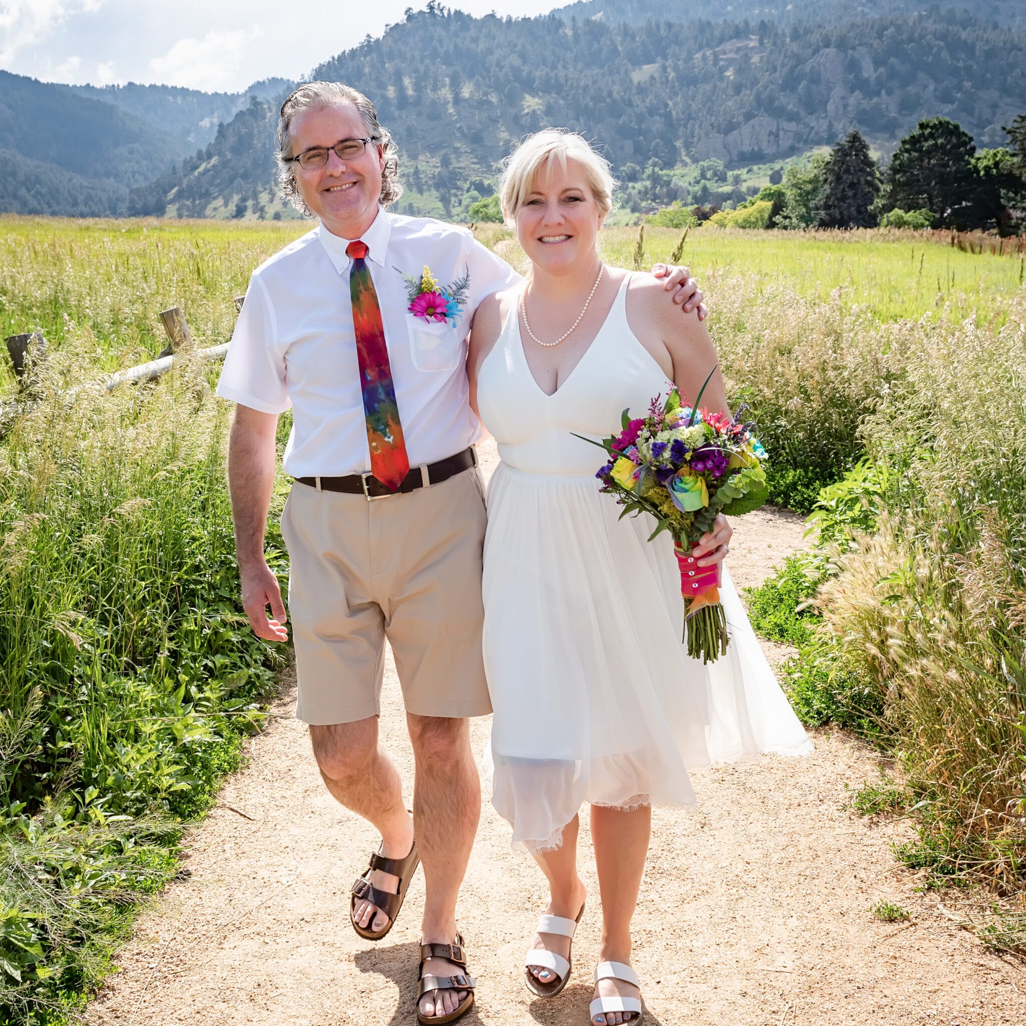 A bride and groom in wedding attire accented with tie-dye elements walk arm in arm on a path at Chautauqua Park.