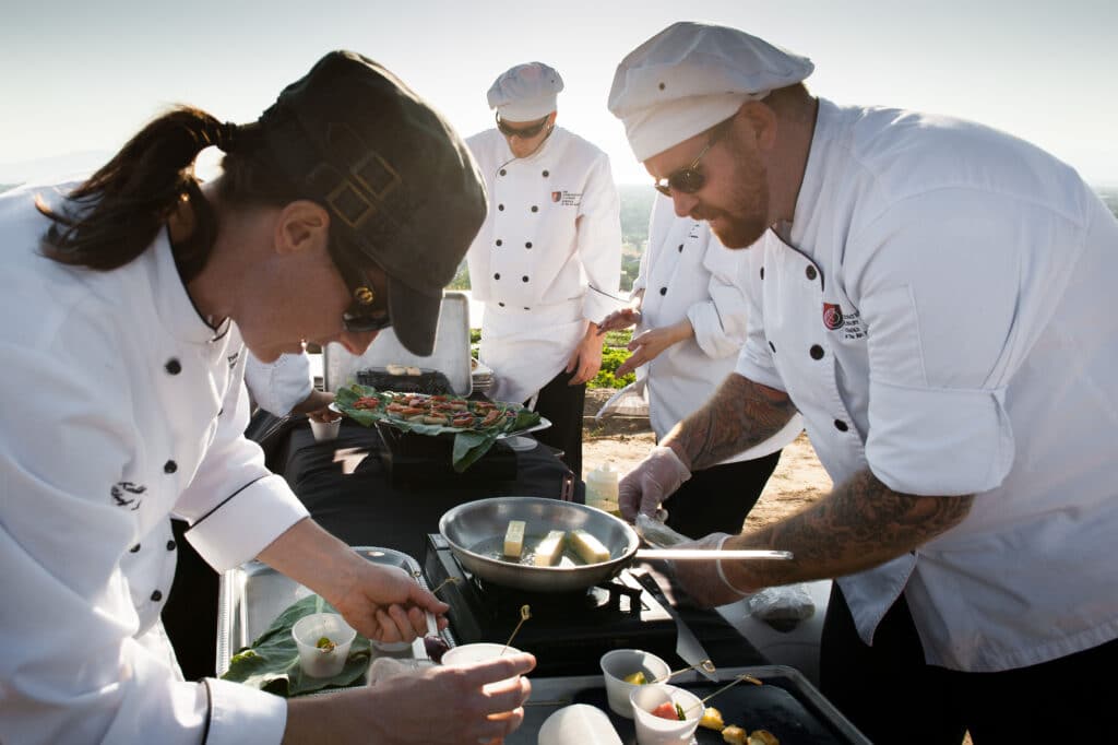 Chef Katie Weinner and several other chefs use portable burners to cook outdoors in the sunshine at a farm pop-up dinner event.
