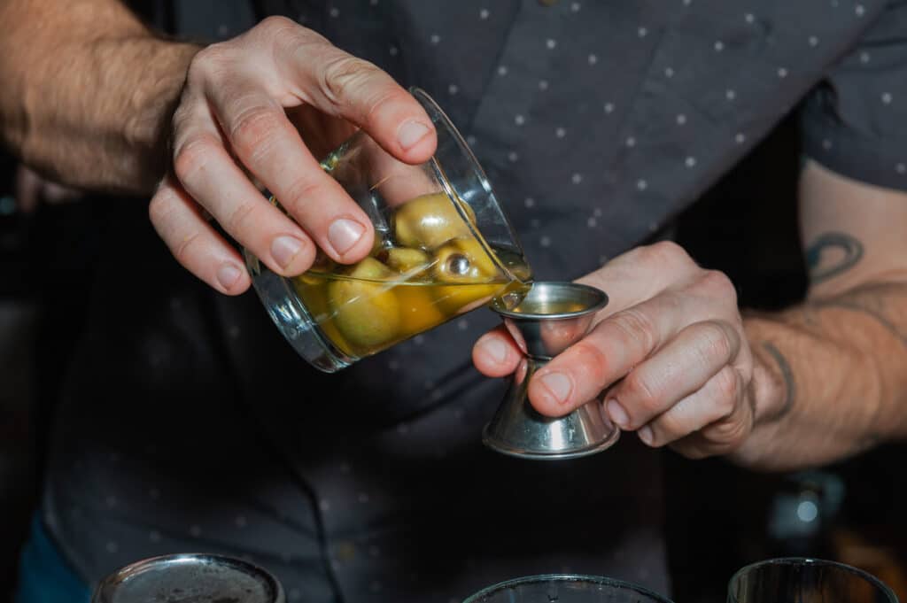 Close up of a mixologist's hands as he pours green olive juice into a jigger while preparing a cocktail.