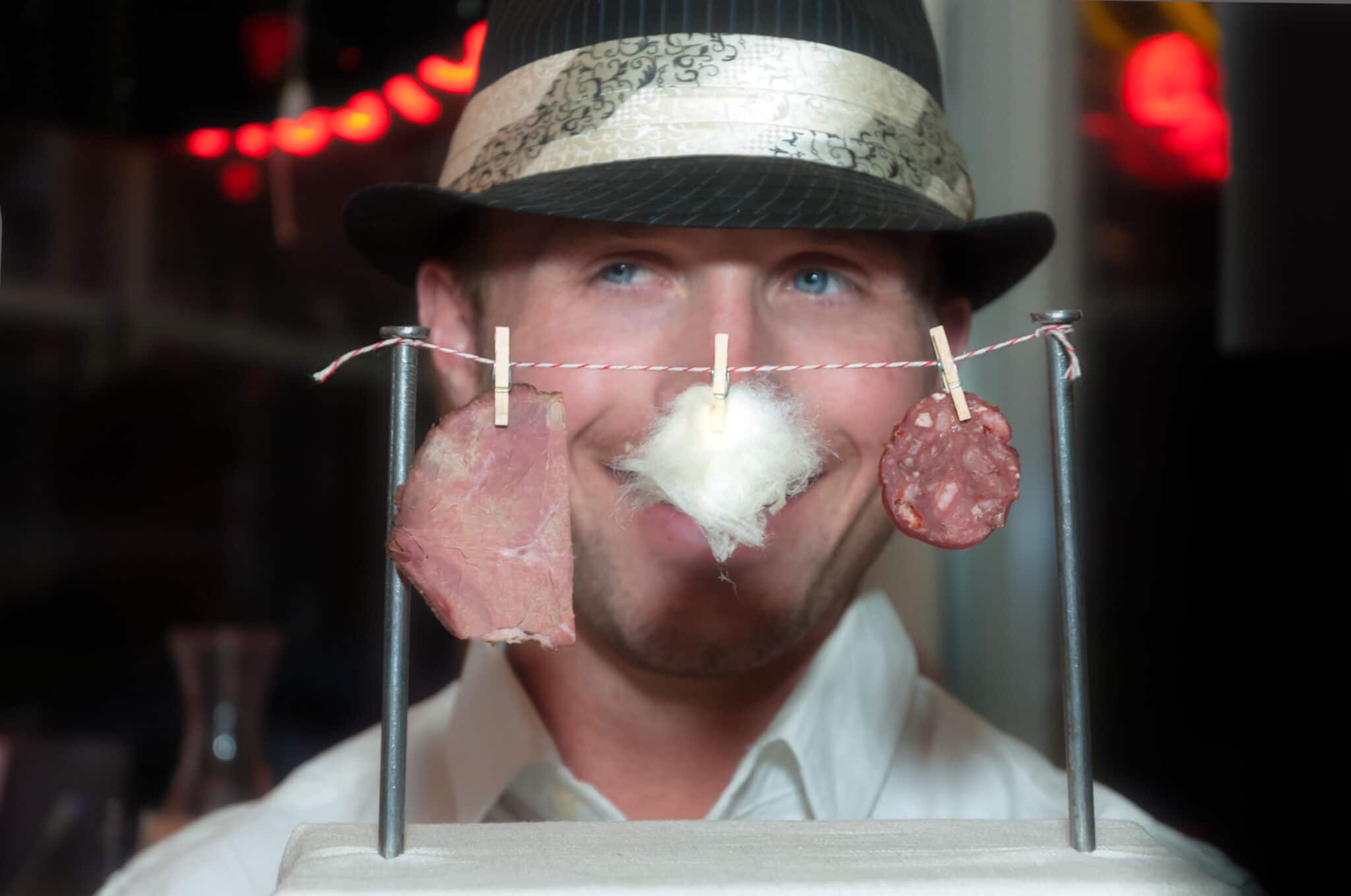 A man wearing a brimmed dress hat playfully places his face behind a clothesline of charcuterie at a pop up restaurant event held by Chef Katie Weinner.