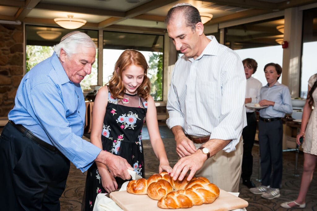 A young girl with her grandfather and father on her side cuts the special bread shaped in the letter M at her bat mitzvah.