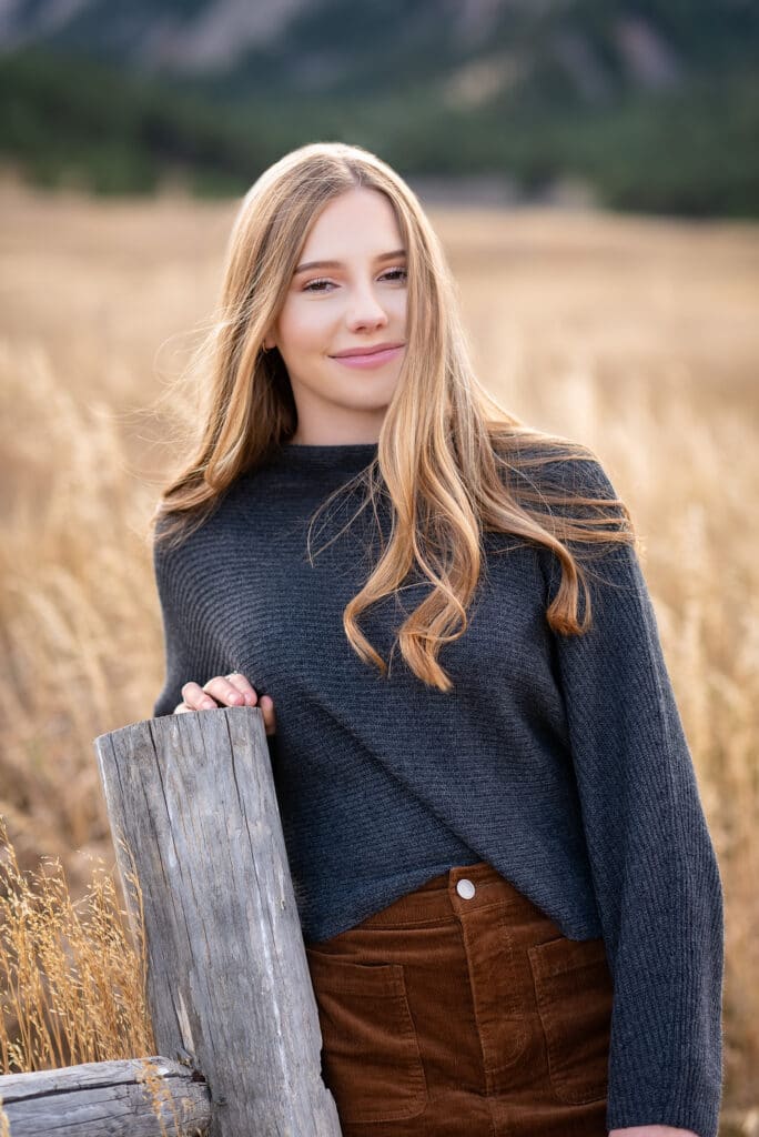 A soon-to-be senior high school graduate woman stands by a split-rail fence post and smiles for her photo.