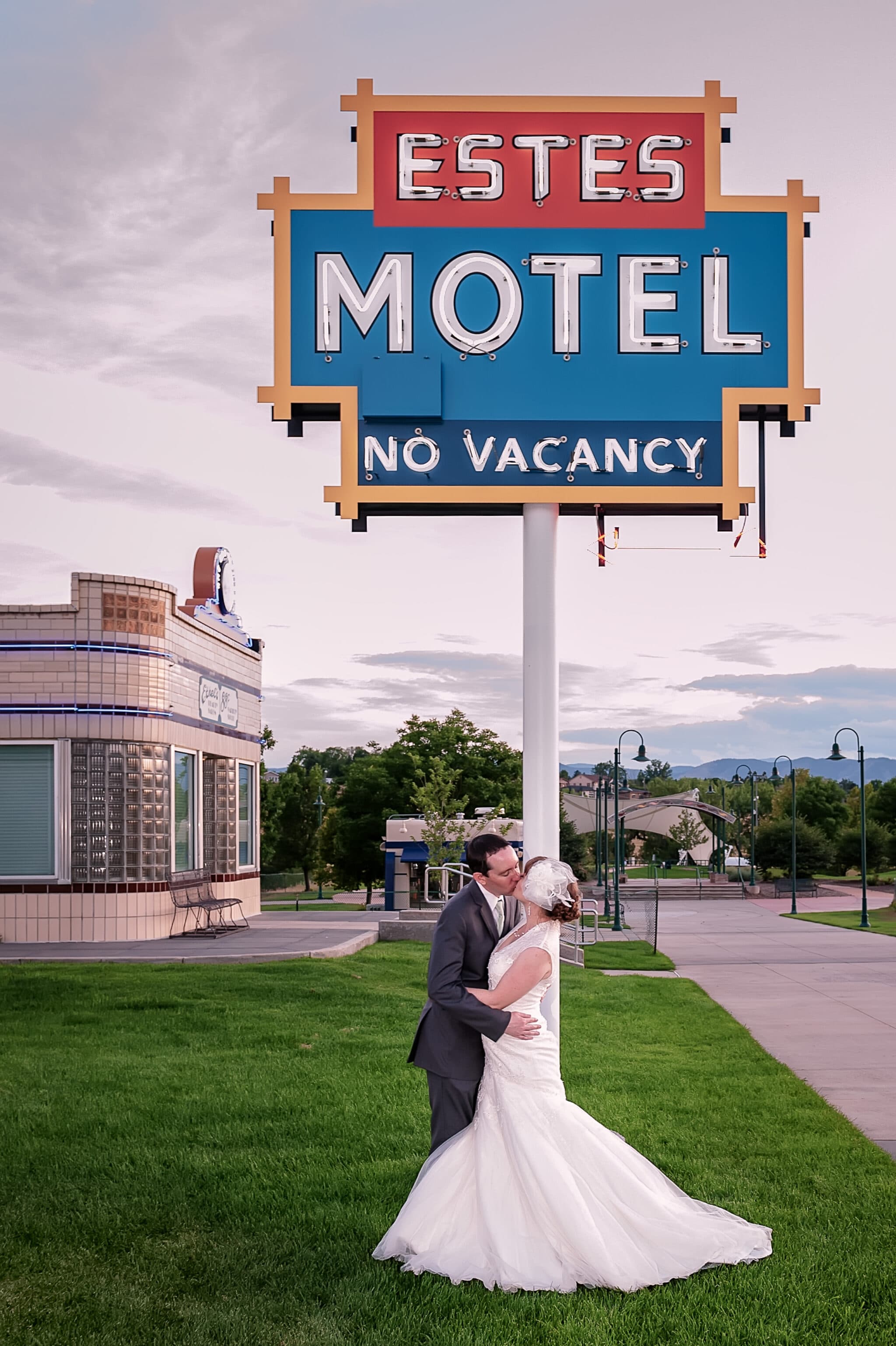 A bride and groom kiss in front of a historic motel sign that says "Estes Motel - No Vacancy" at Lakewood Heritage Center.